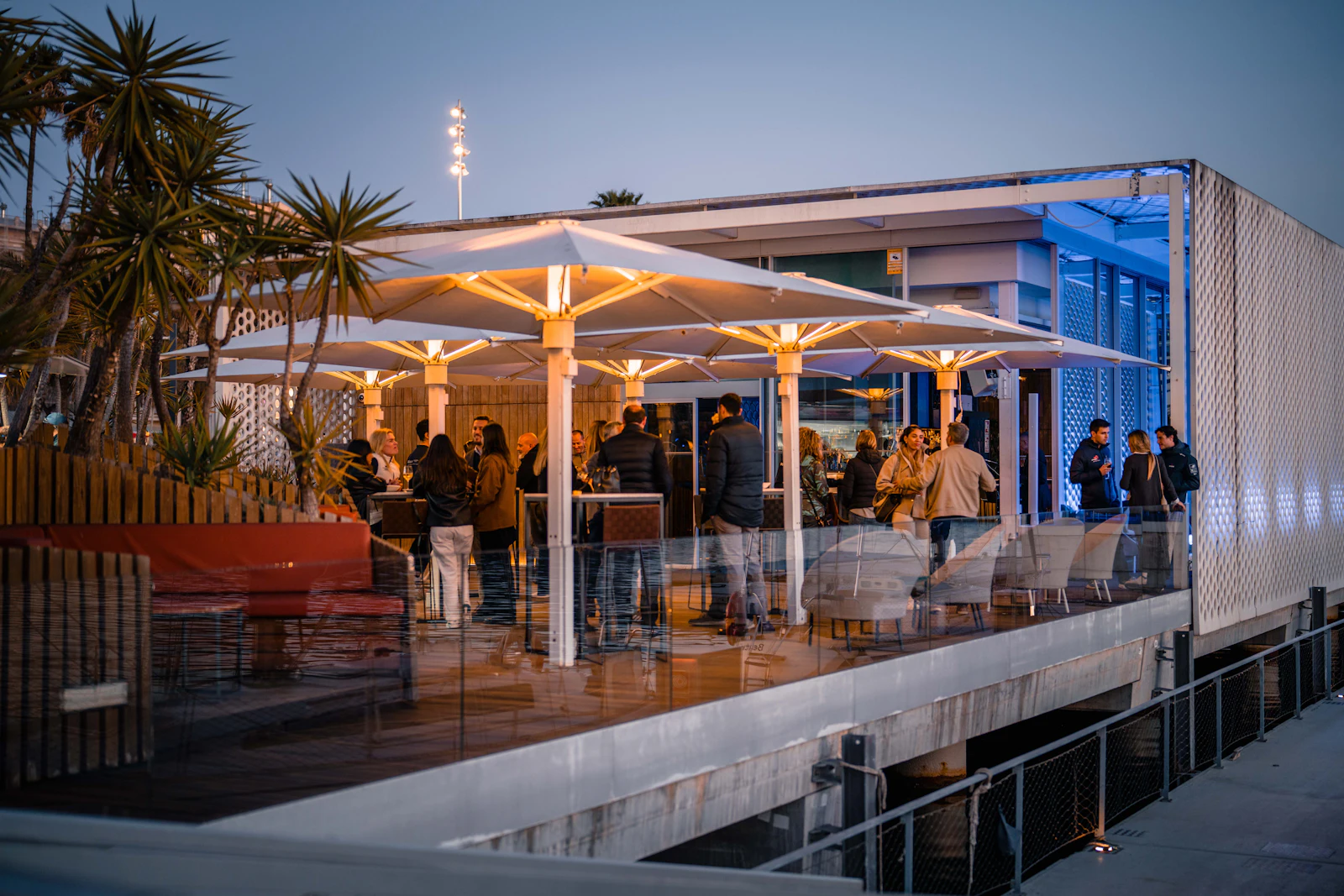 Waterfront pavilion with illuminated umbrellas and guests gathered at dusk in Barcelona marina.