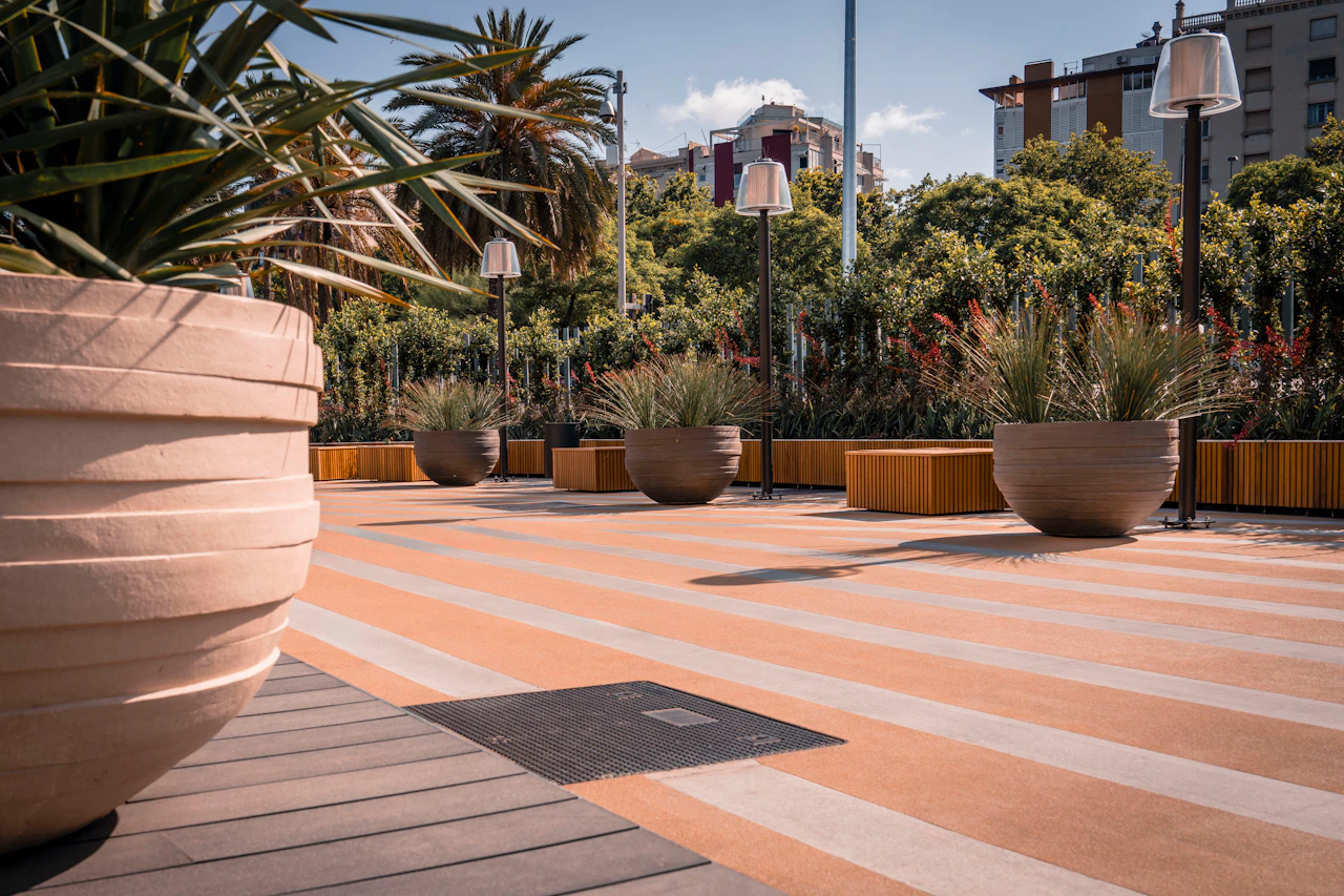 Waterfront promenade with terracotta paving, large planters, palm trees, and modern street lamps overlooking Barcelona buildings.
