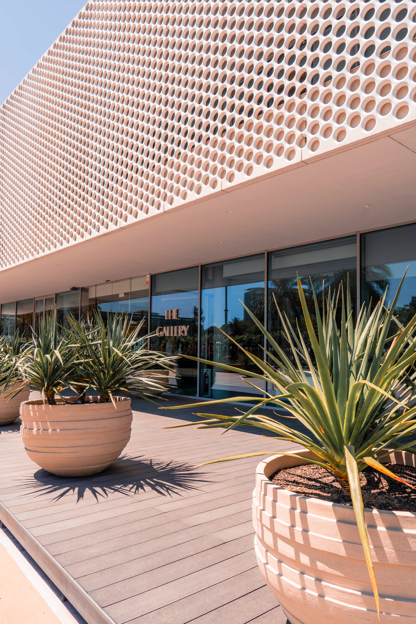 Edificio moderno frente al mar con fachada perforada y plantas en macetas sobre una cubierta de madera.