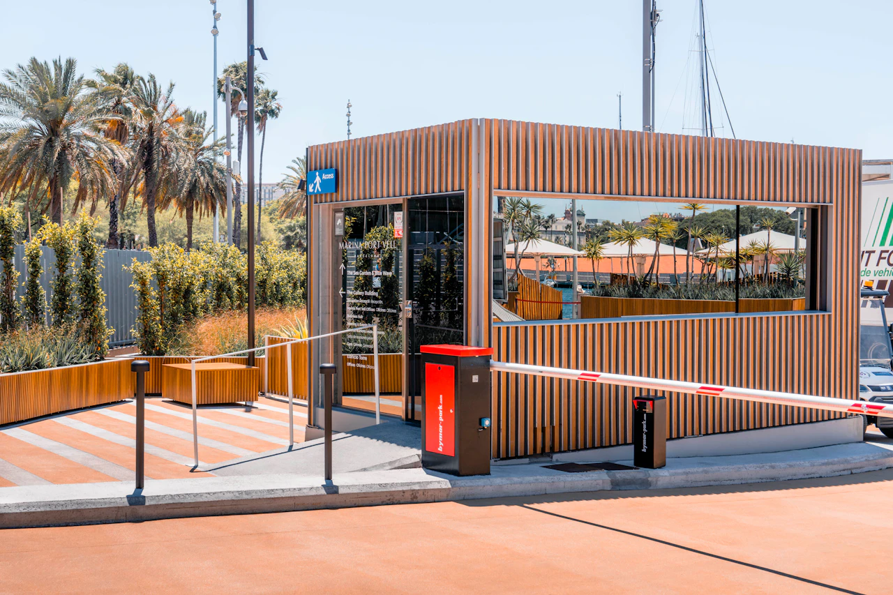 Modern corrugated metal kiosk with orange accents at a marina waterfront with palm trees and sailboats.