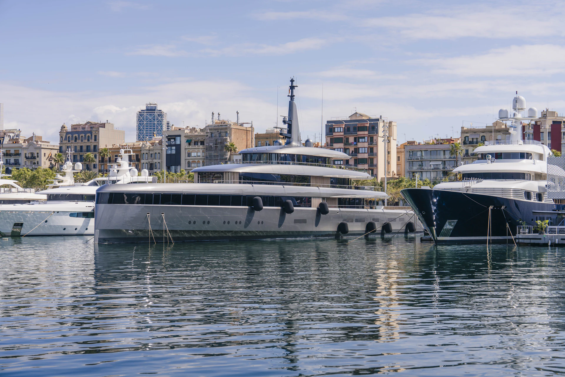 Varios superyates amarrados en Marina Port Vell con el paisaje urbano y los edificios de Barcelona al fondo.