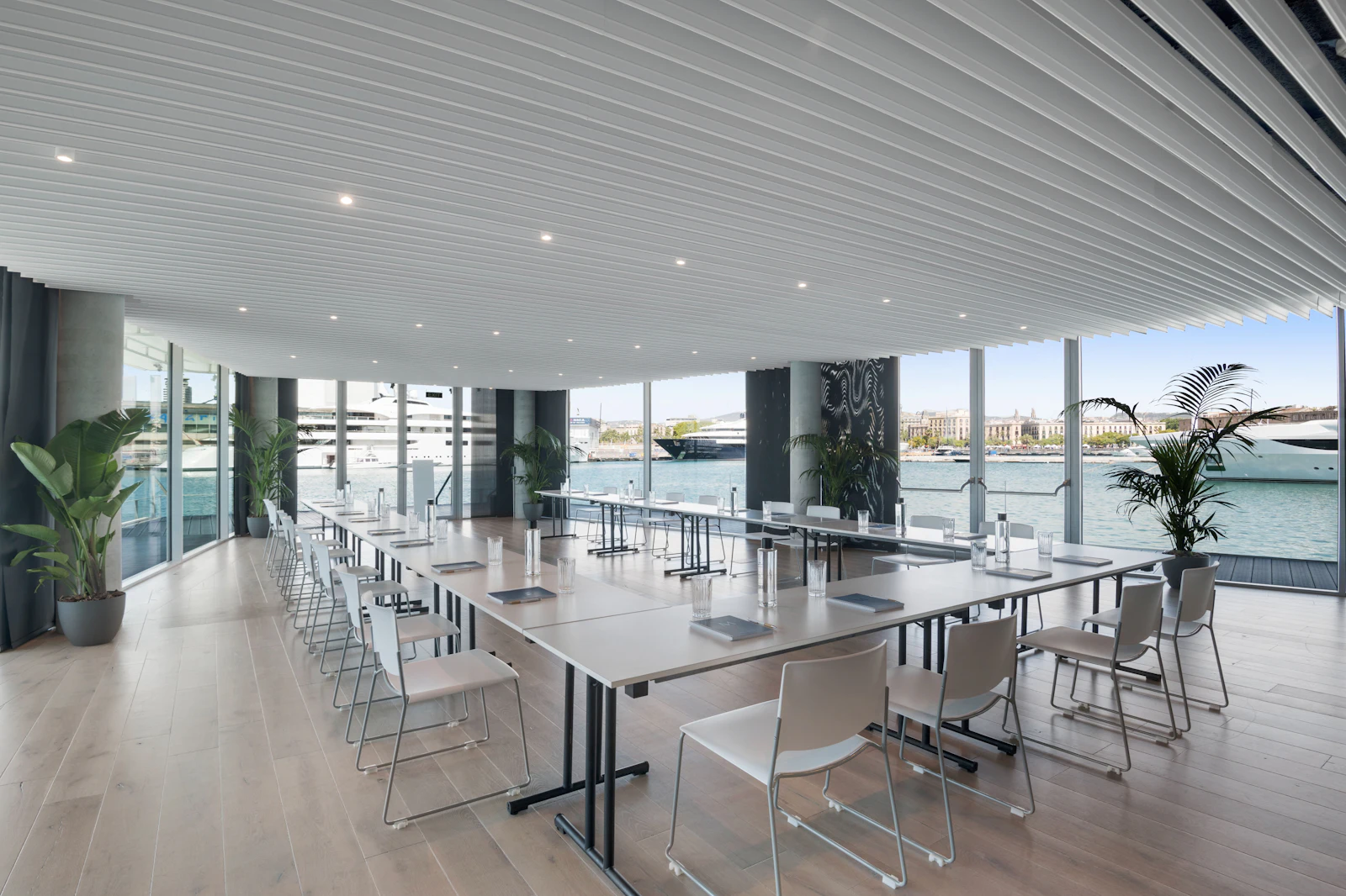 Espacio moderno para eventos con mesas y sillas blancas y ventanales del suelo al techo con vistas al puerto de Barcelona y a los superyates amarrados.