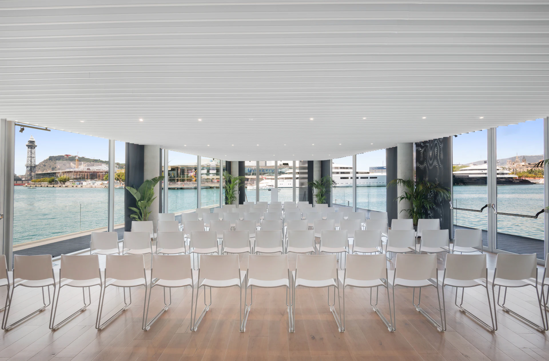 Sala de conferencias moderna con sillas blancas dispuestas en filas con vistas al frente marítimo y la marina de Barcelona.