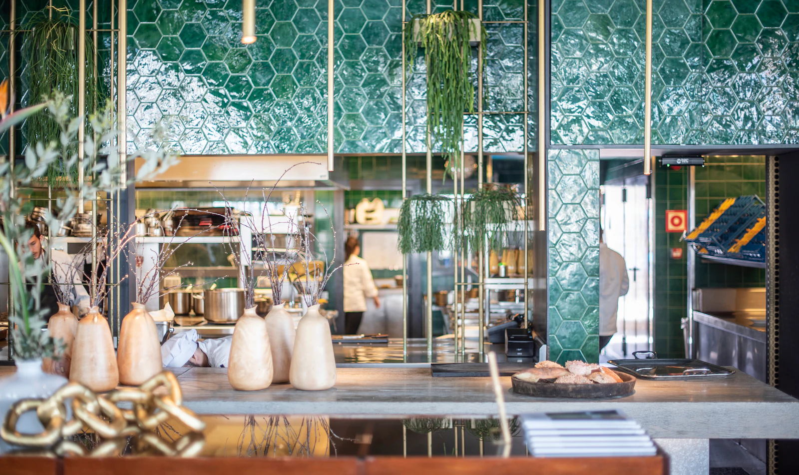 Modern waterfront restaurant with green hexagonal wall panels, hanging plants, and stone decorative vessels on the counter.