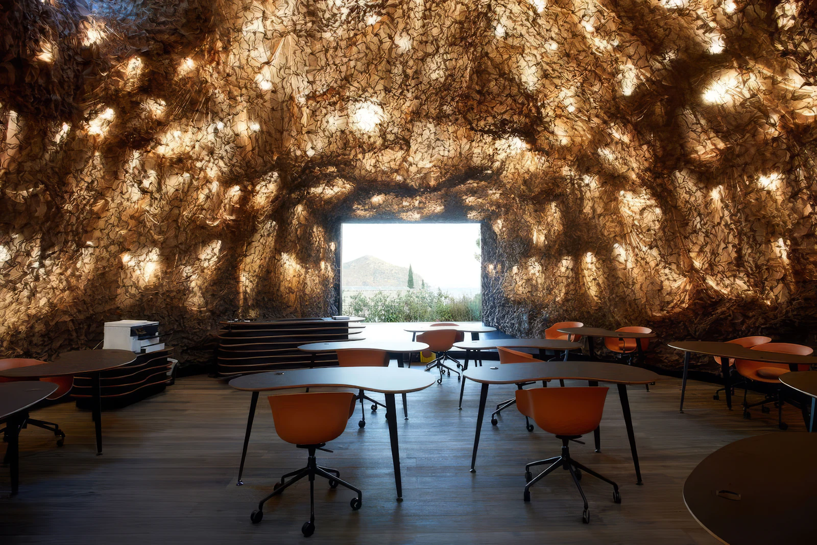 Cave-like room with textured walls, orange chairs and tables, and a window overlooking hills.