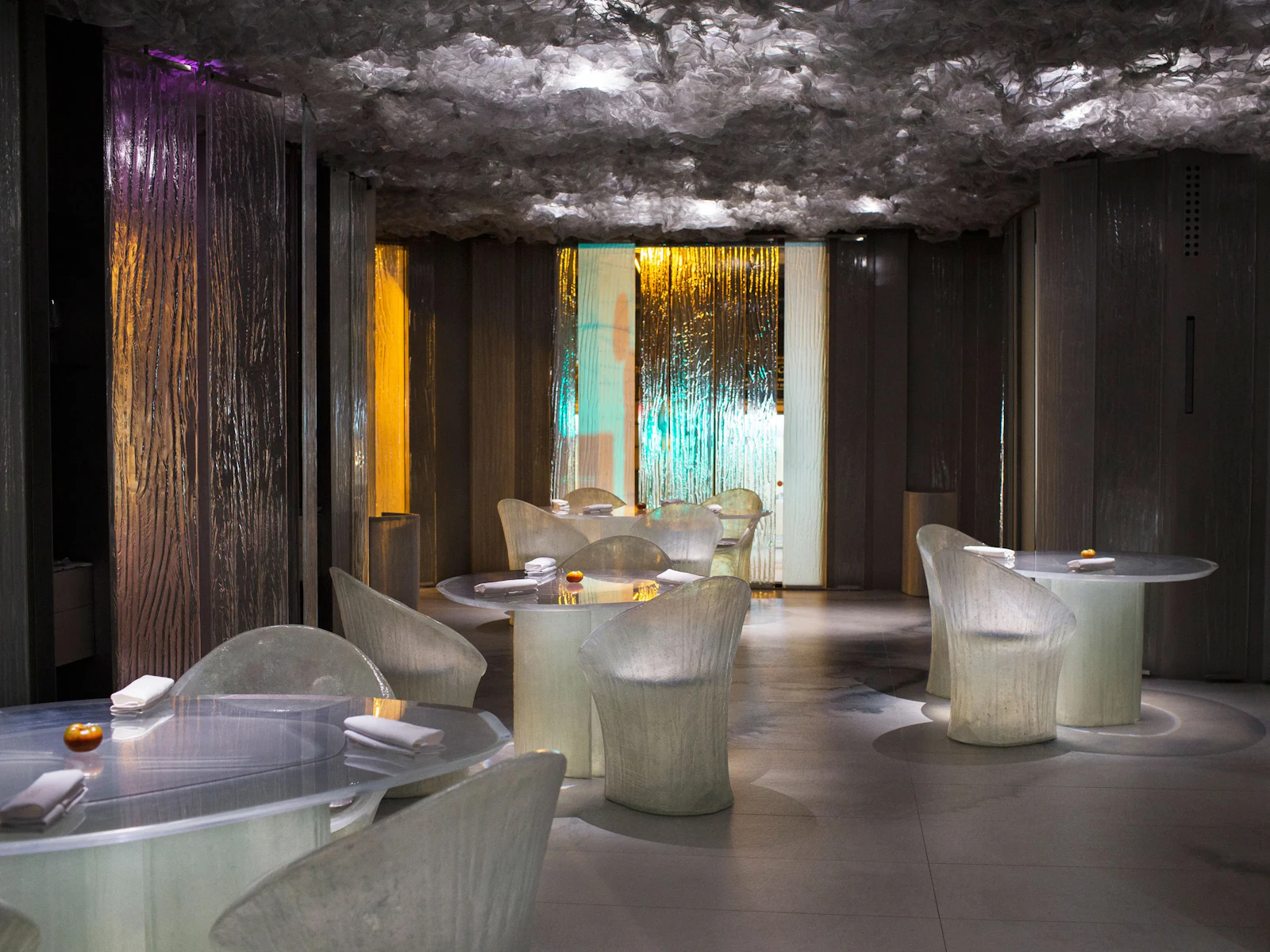 Modern restaurant interior with translucent chairs, round tables and a textured ceiling.