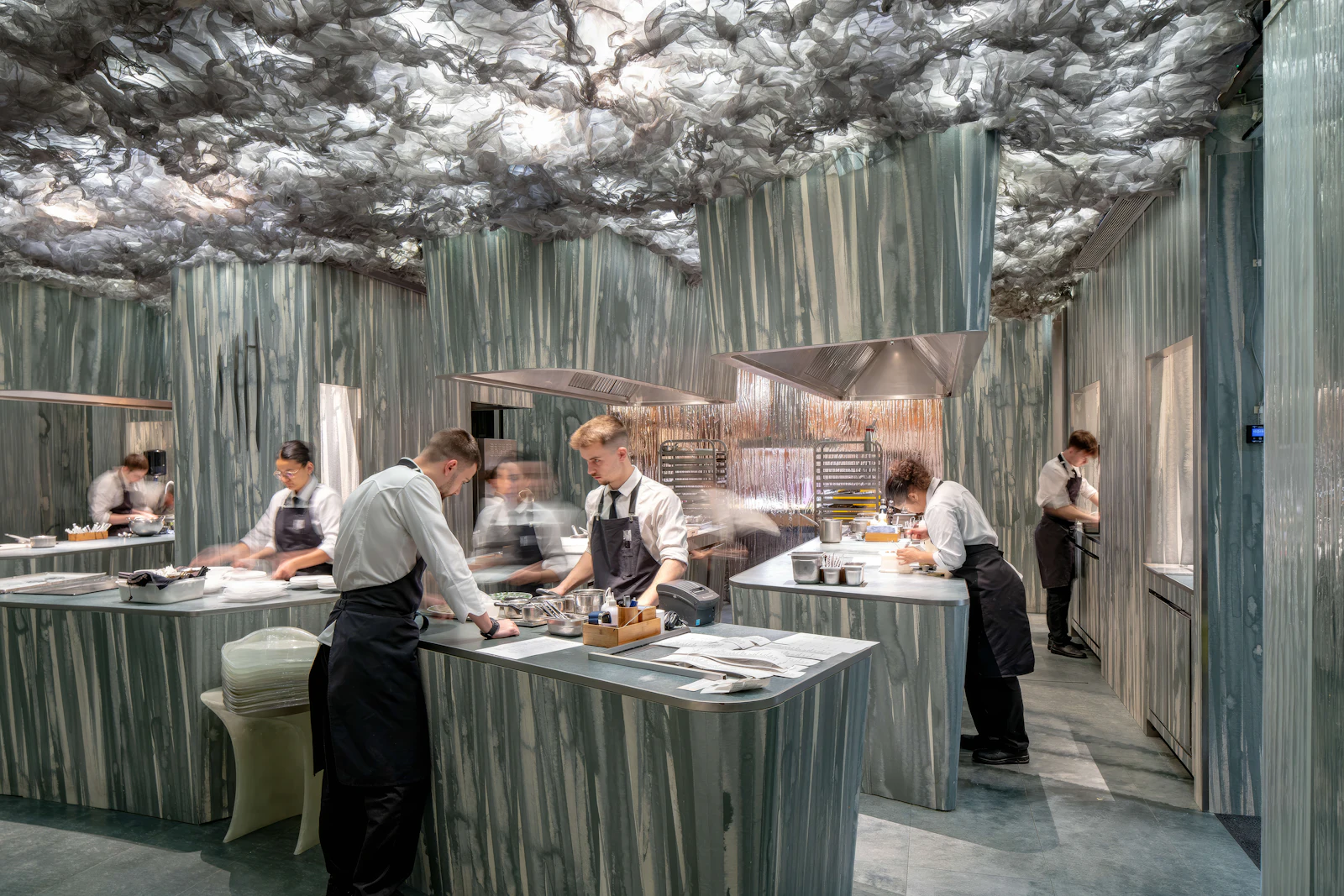 Chefs in aprons work at counters in a marble-clad open kitchen with a sculptural ceiling.