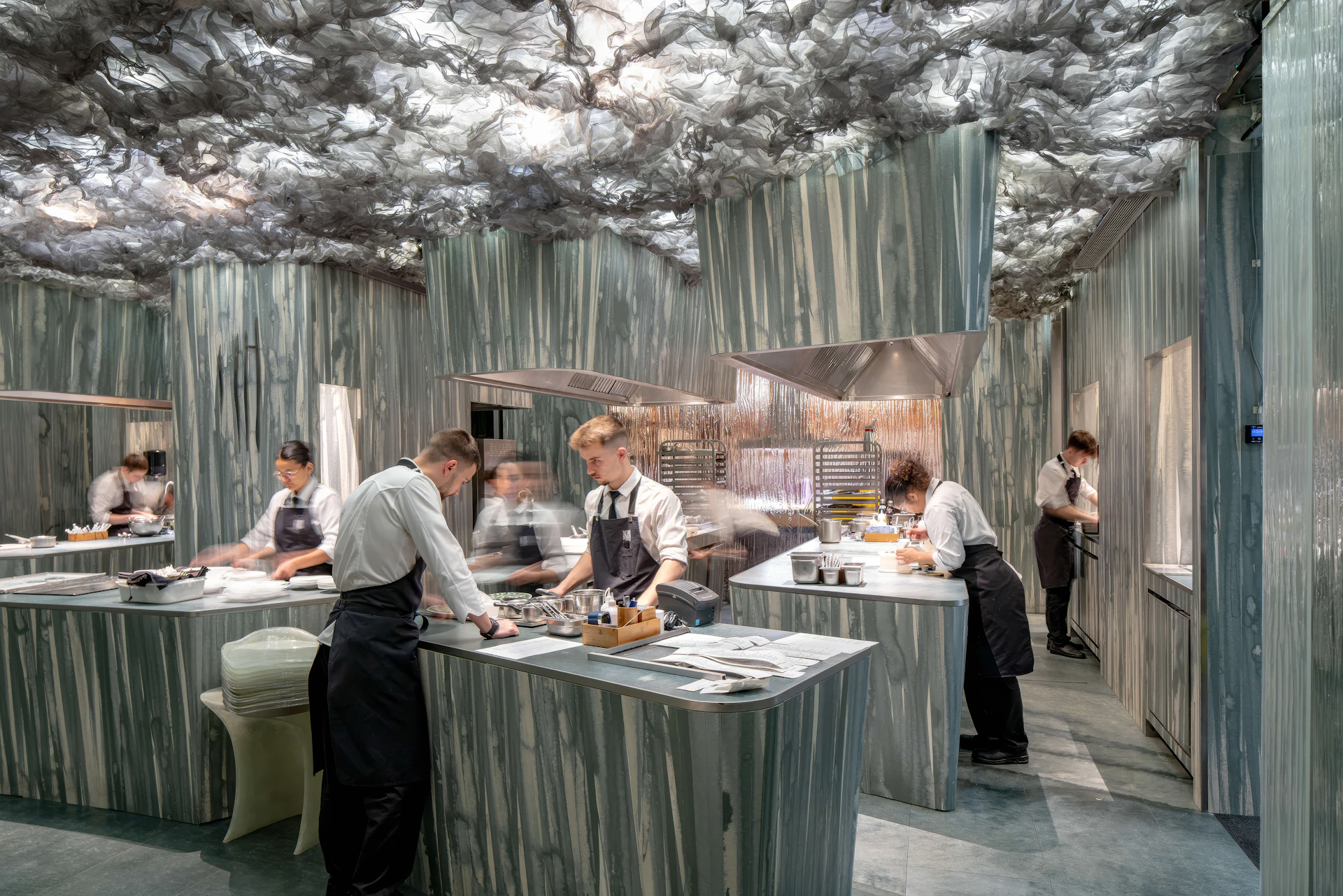 Chefs in aprons work at counters in a marble-clad open kitchen with a sculptural ceiling.