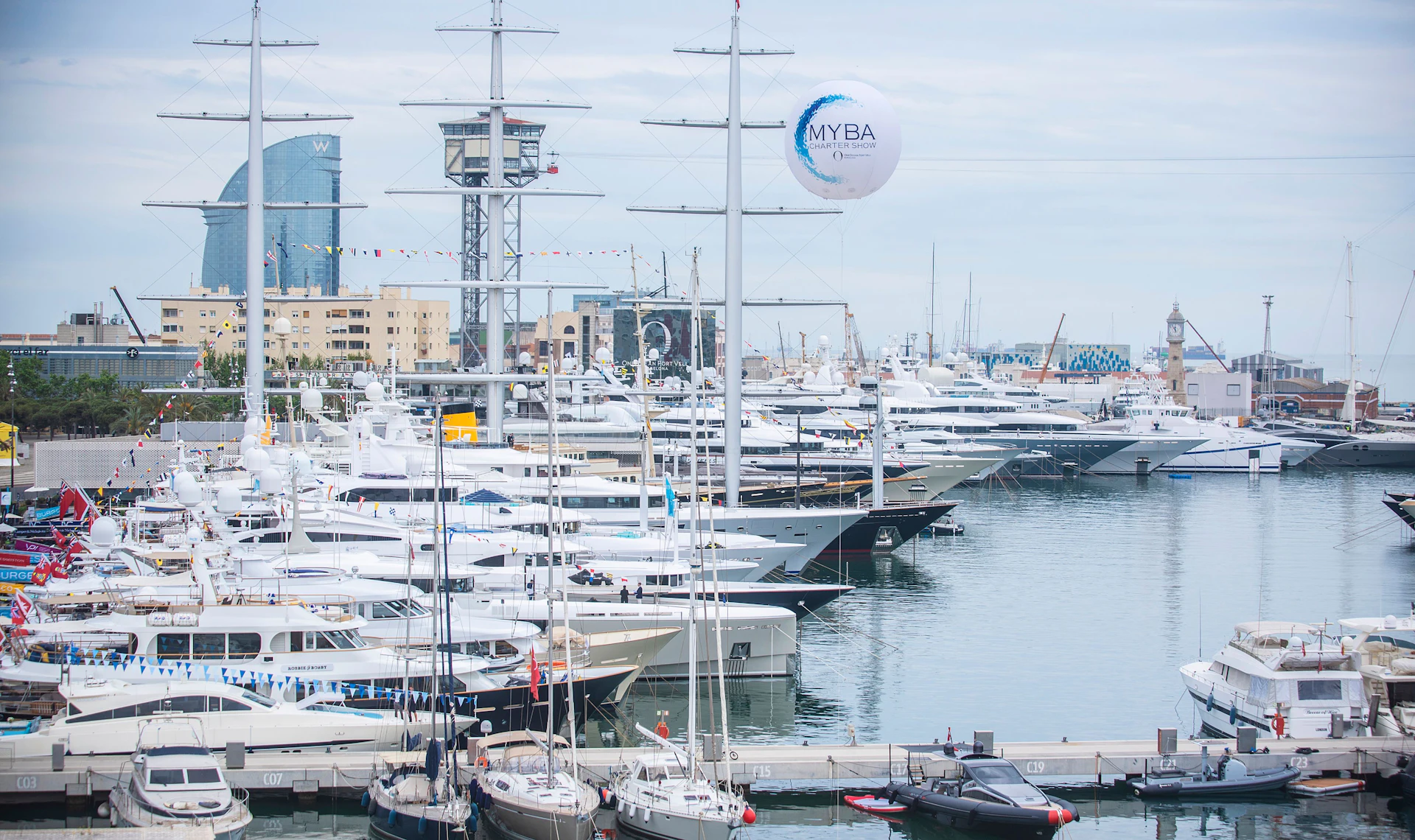 Varios superyates amarrados en Marina Port Vell de Barcelona con el perfil de la ciudad y el globo de MYBA a la vista.