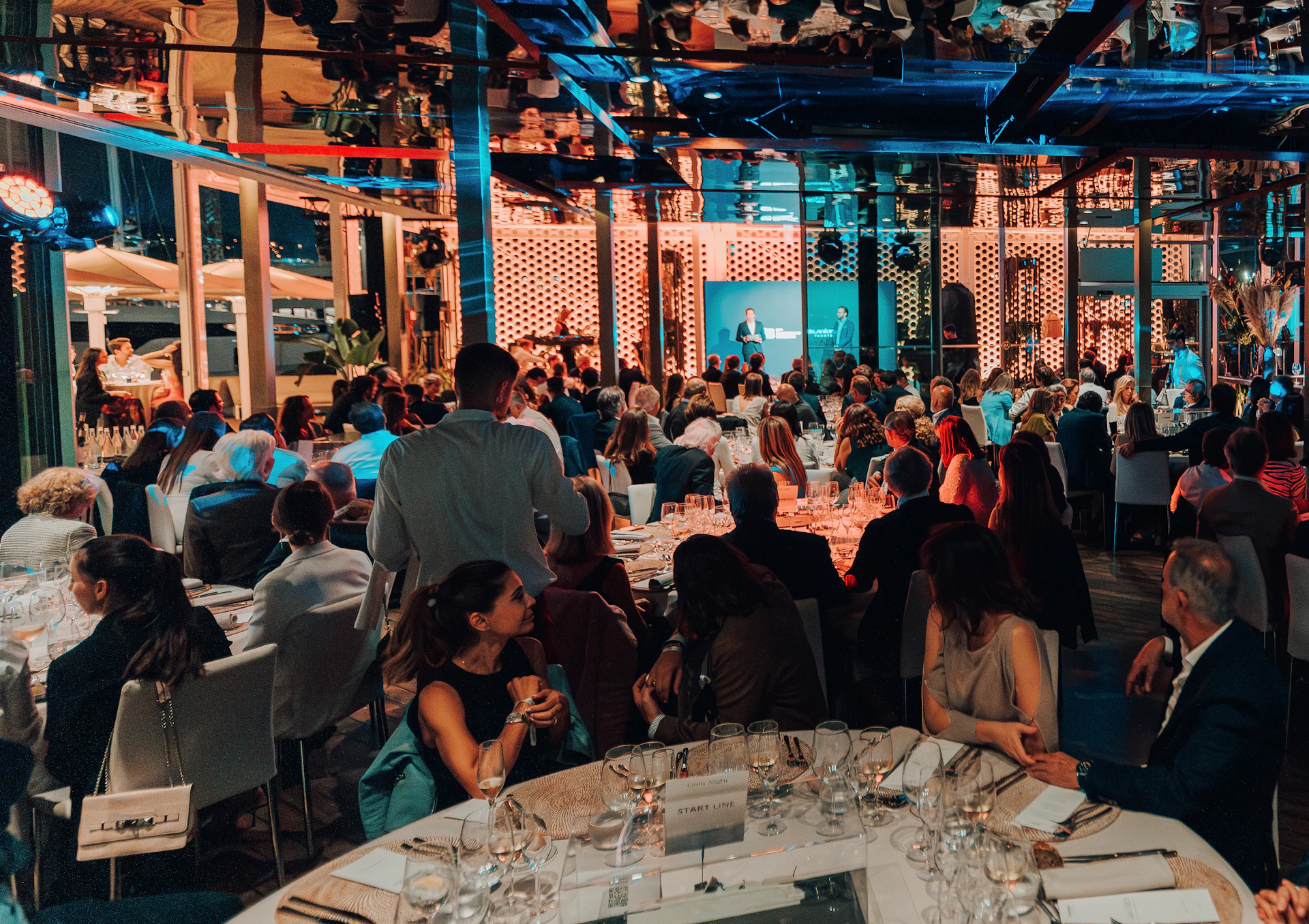 Guests dining at tables inside an illuminated waterfront venue with floor-to-ceiling windows overlooking Barcelona's harbor.