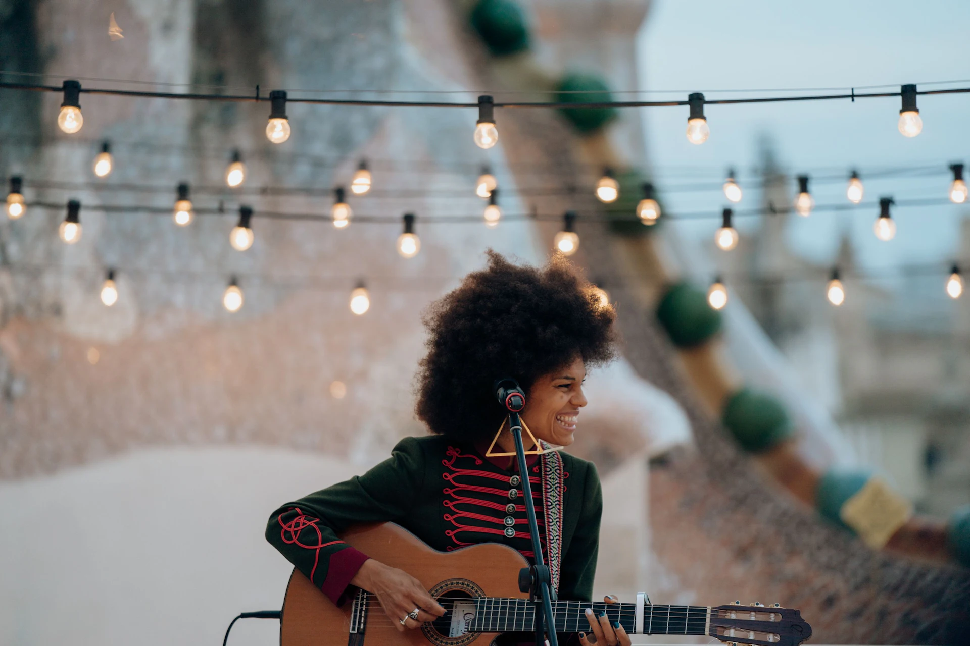 Mujer tocando la guitarra acústica bajo guirnaldas de luces en un espacio al aire libre con arquitectura histórica.