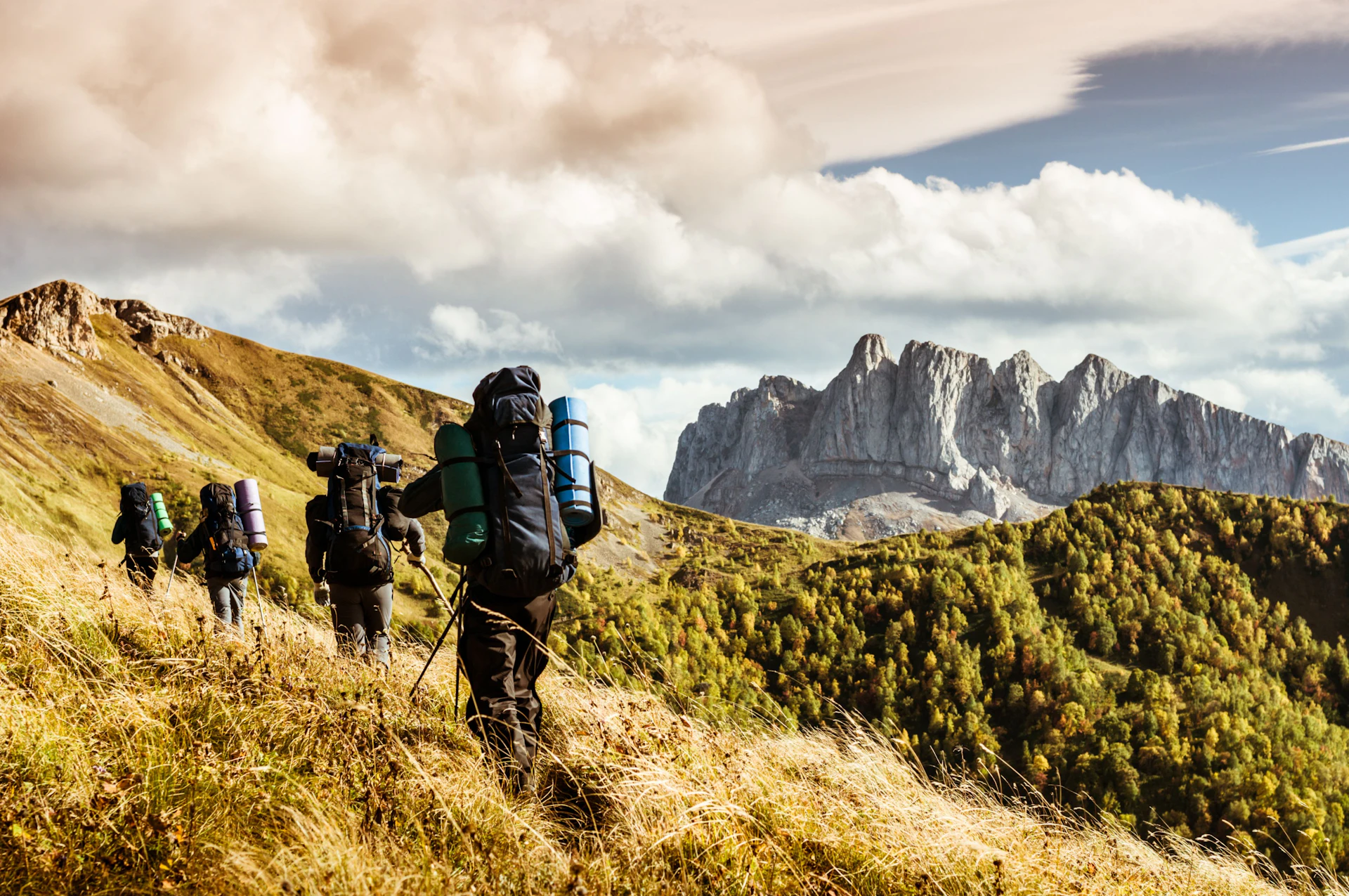 Excursionistas con grandes mochilas recorren un sendero de montaña con espectaculares picos rocosos a lo lejos.