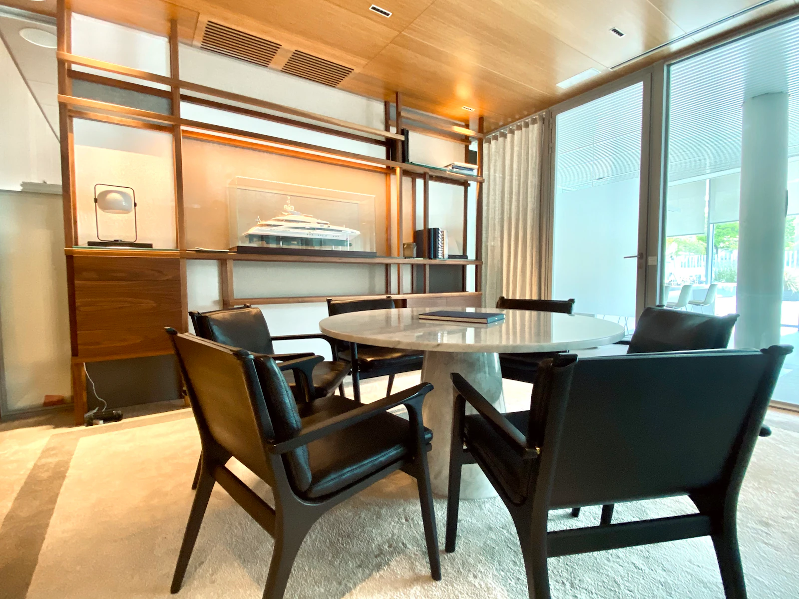 Modern office or lounge space with round table, black chairs, wooden shelving, and water views through glass doors.
