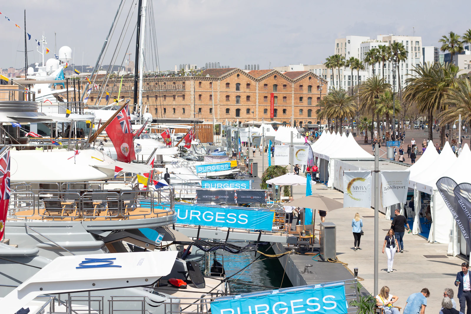 Superyachts moored at Barcelona's Port Vell with red brick warehouse buildings and palm trees in background.