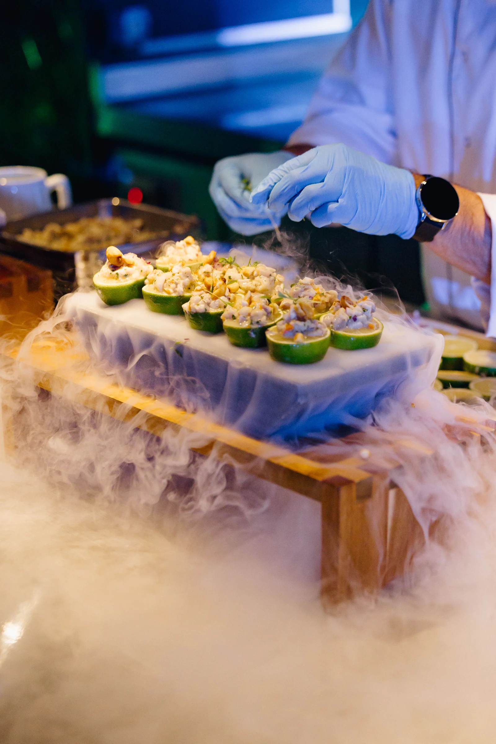 Chef con guantes decorando mitades de lima rellenas de marisco en una mesa de presentación con hielo seco.