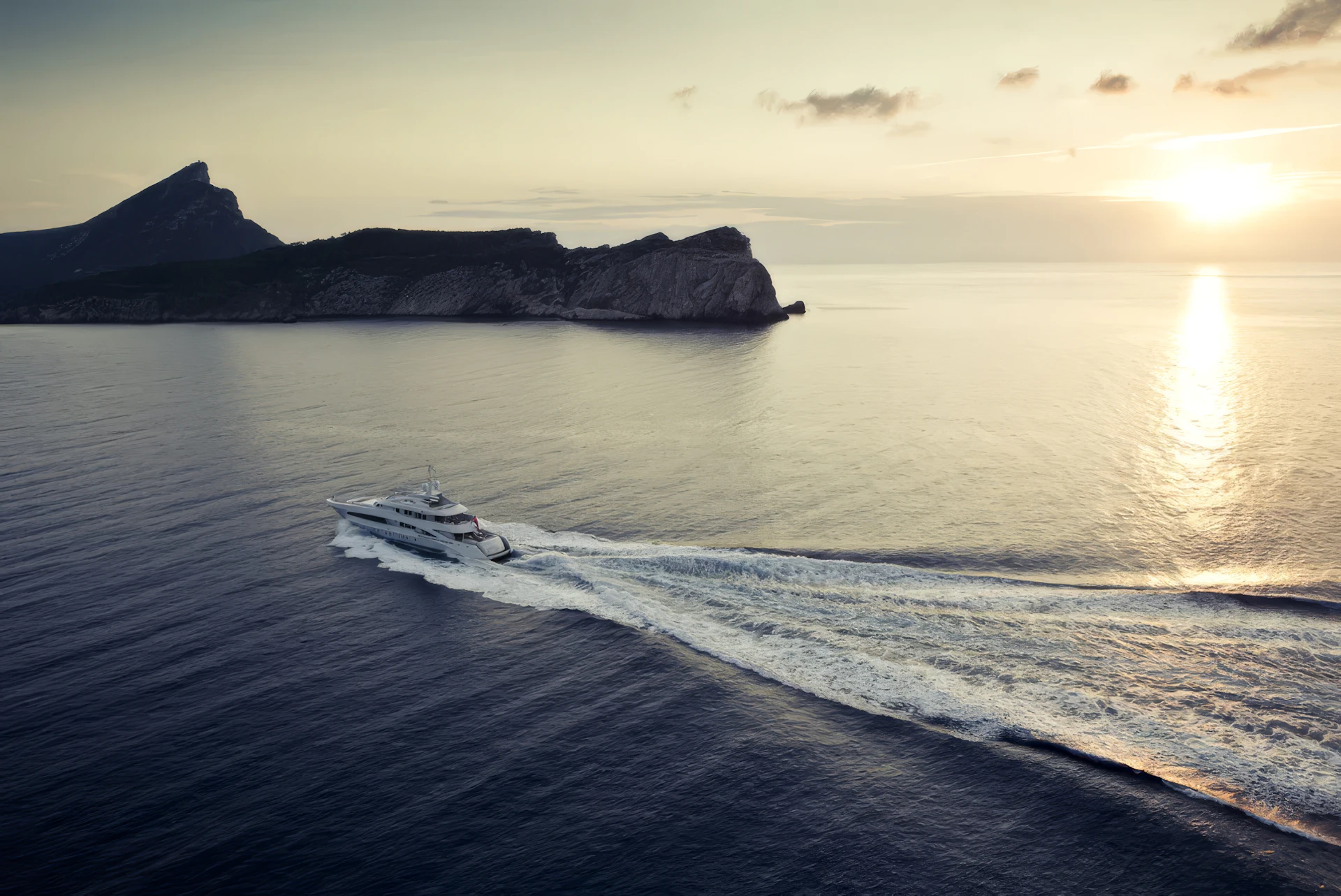 Superyacht moving through calm waters at sunset with rocky island and coastline in background.