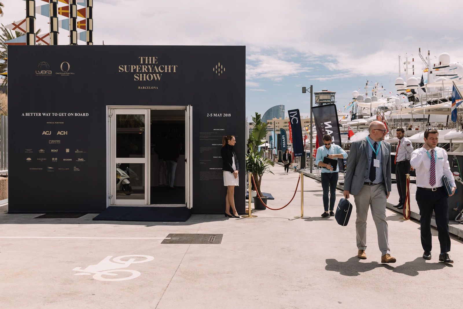 The Superyacht Show exhibition booth at Barcelona marina with visitors and moored yachts in background.
