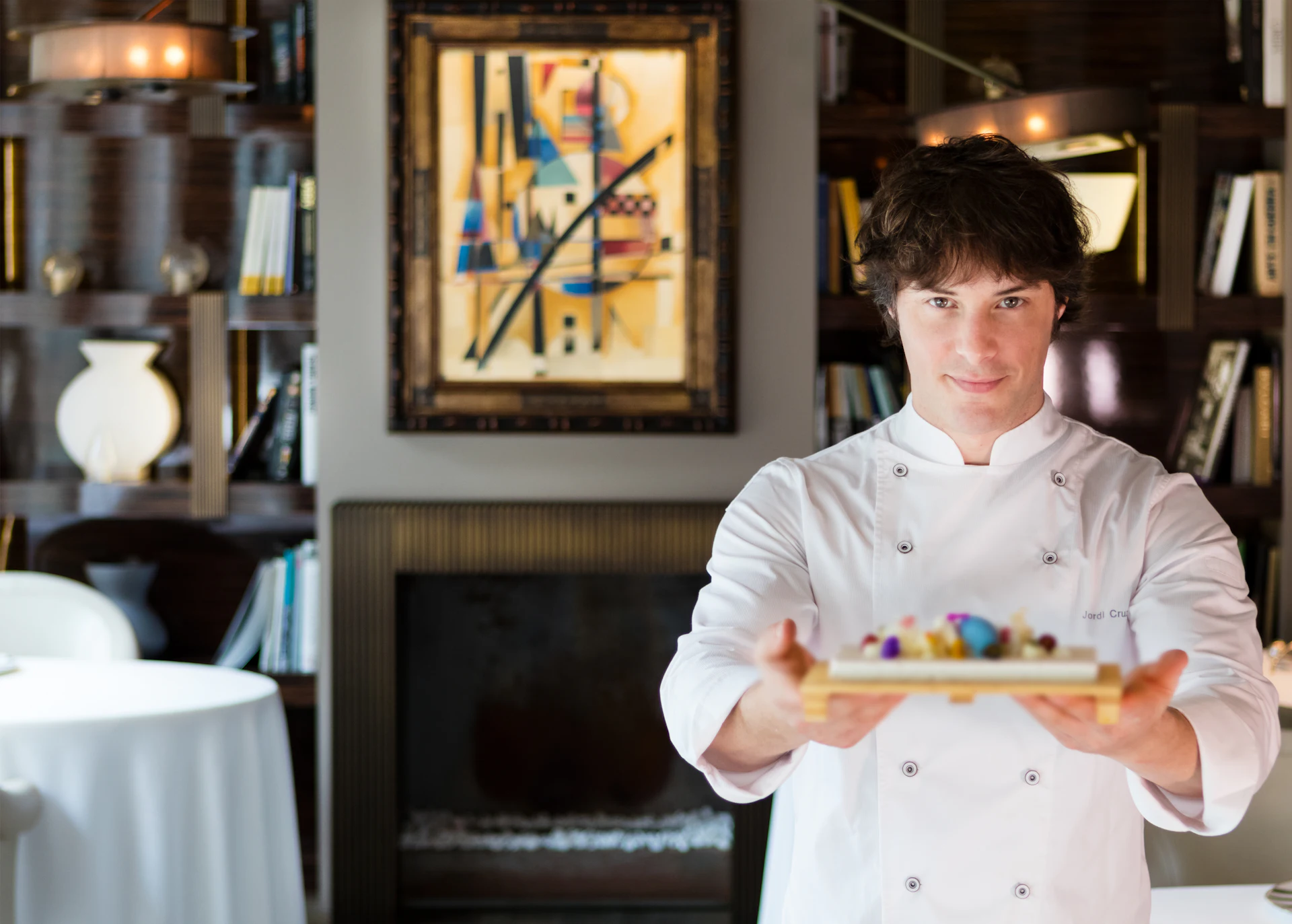 Chef in white chef's coat presenting a plated dessert in an elegant dining room with bookshelves and artwork.
