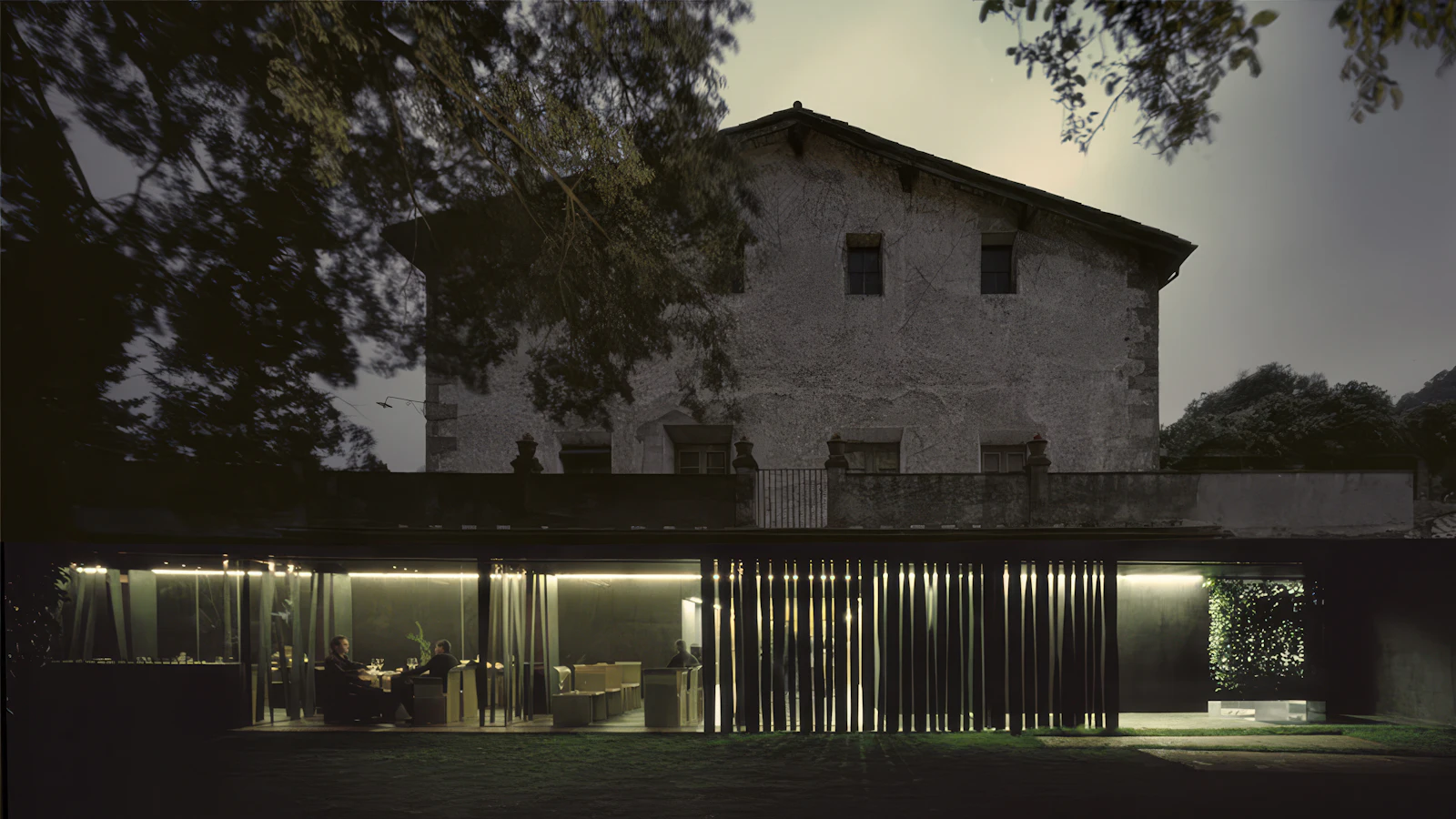 Diners seated in a lit modern glass extension beneath an old stone building at dusk.