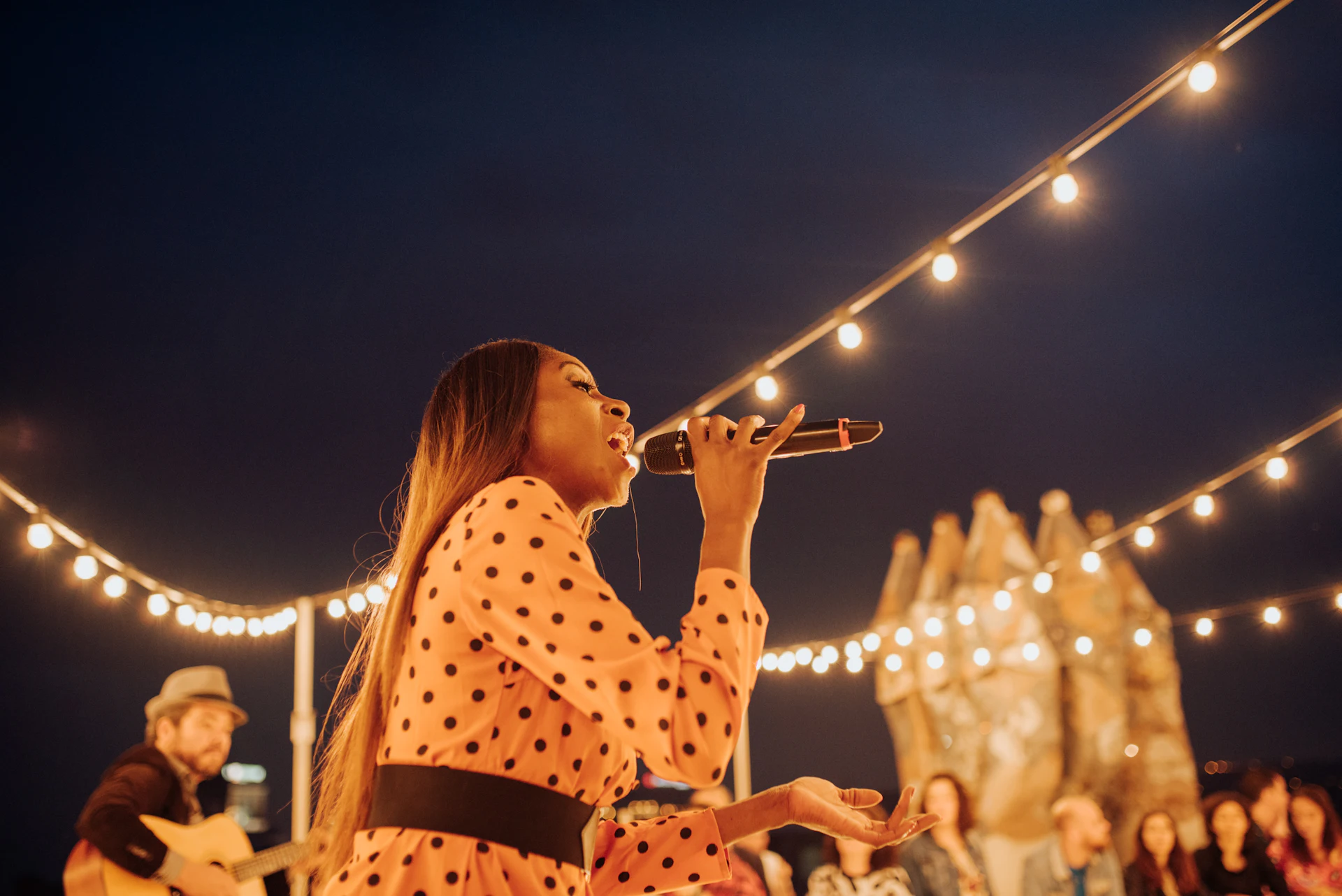 Female singer performing with microphone under string lights at an evening event.