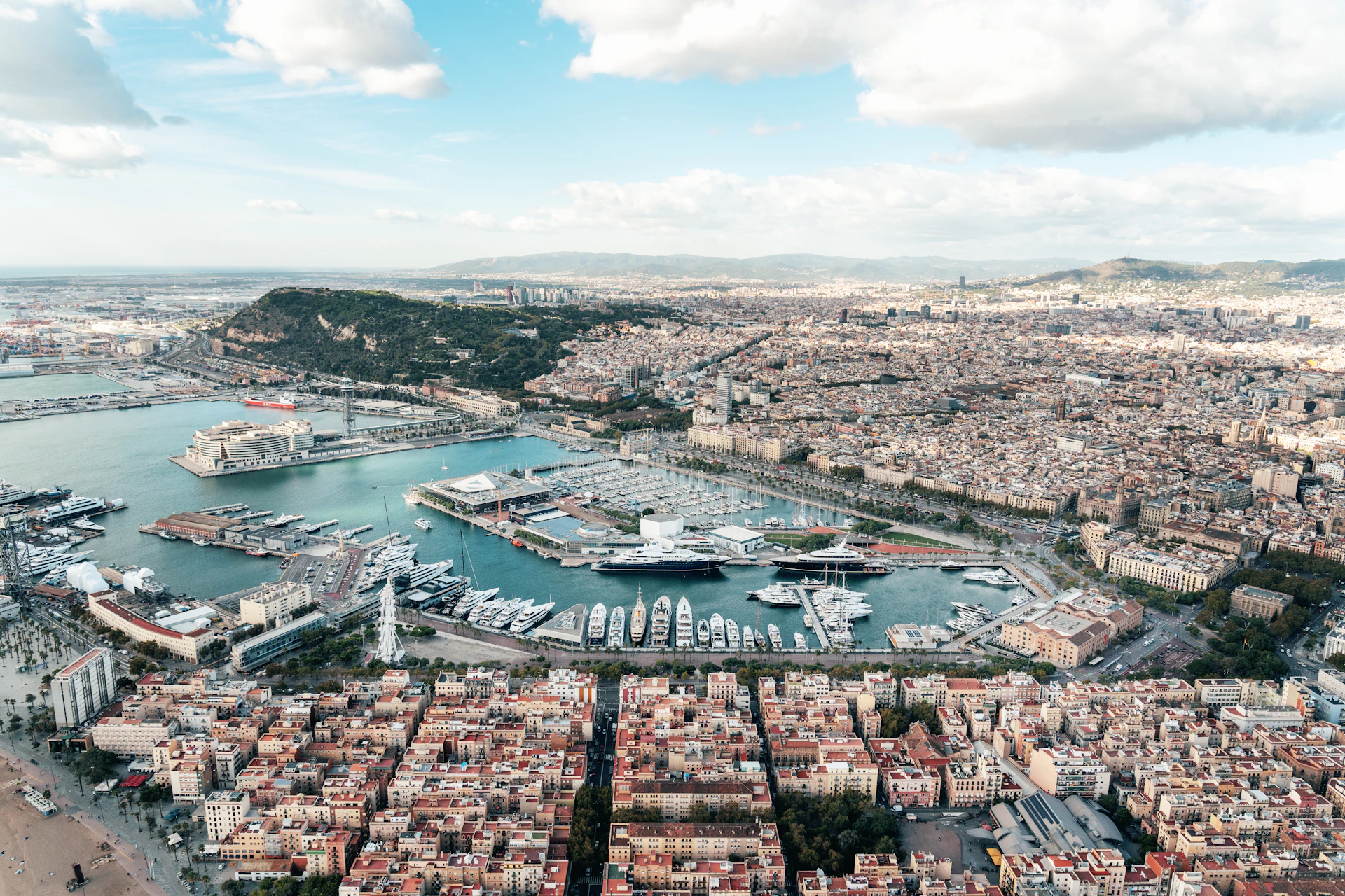 Aerial view of Barcelona marina with moored superyachts and city extending toward distant hills.