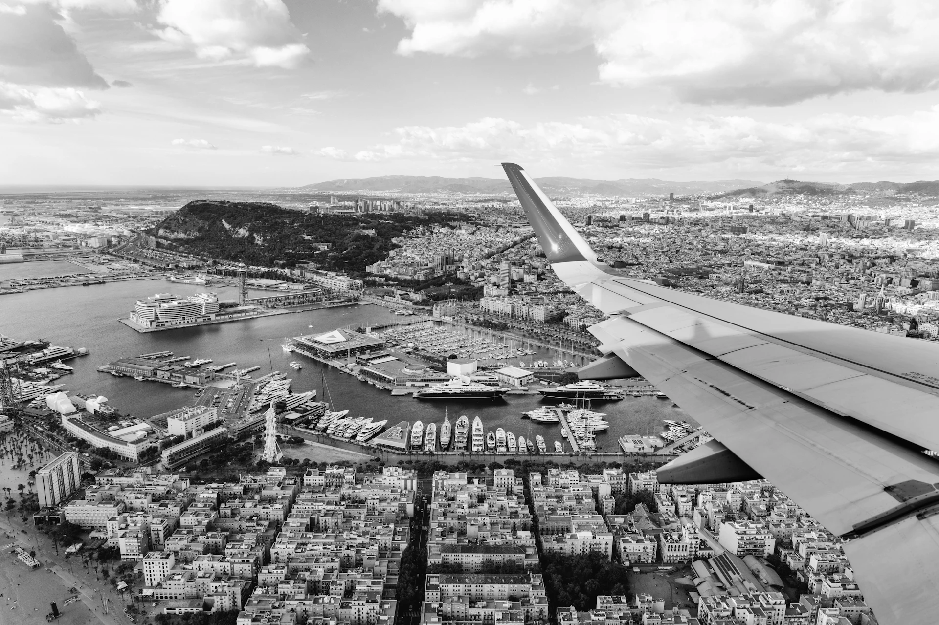 Ala de avión en vuelo sobre el puerto deportivo de Barcelona con superyates amarrados y el frente marítimo de la ciudad visibles abajo.