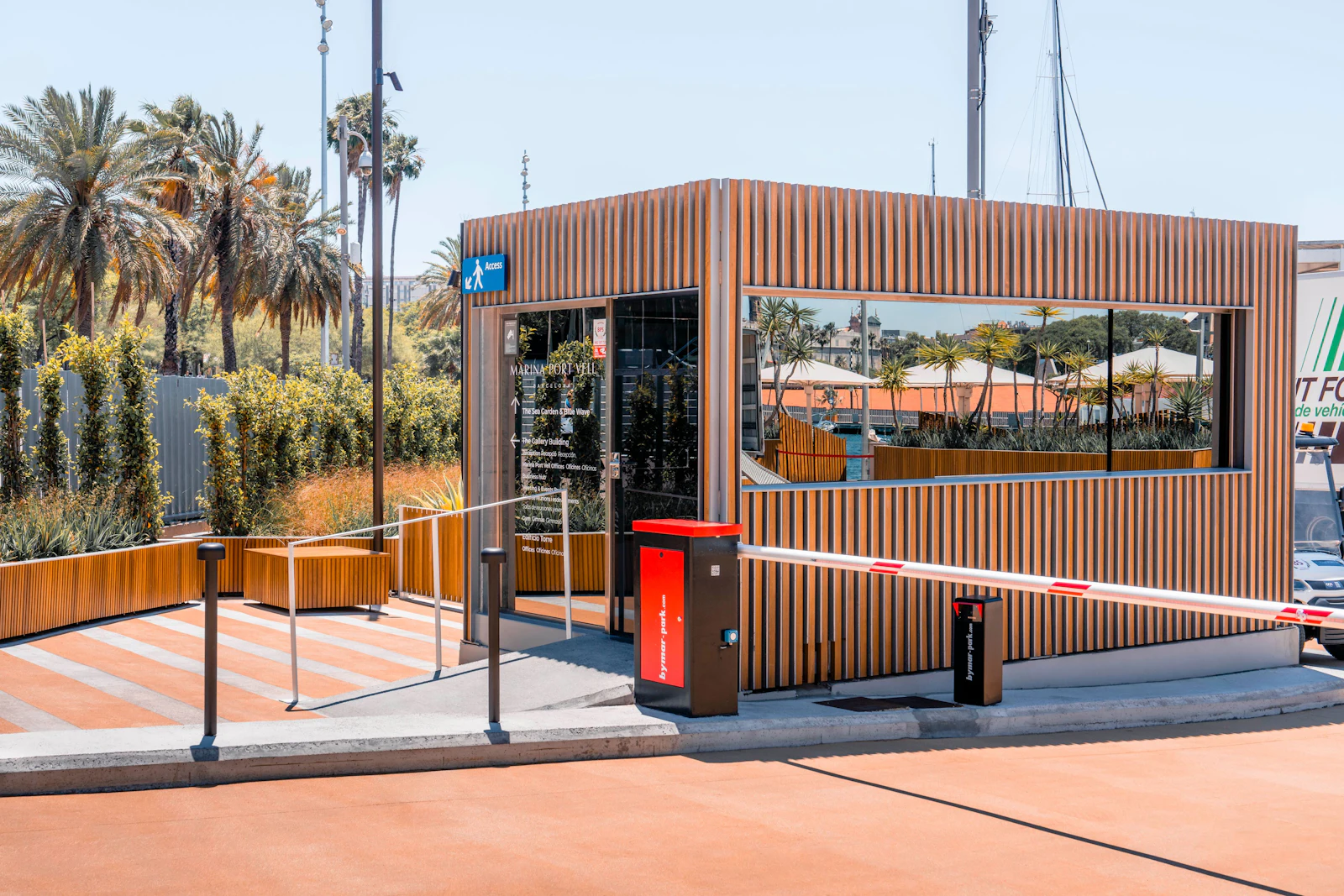 Moderna caseta de madera con detalles rojos en la entrada de la marina, rodeada de palmeras y yates.