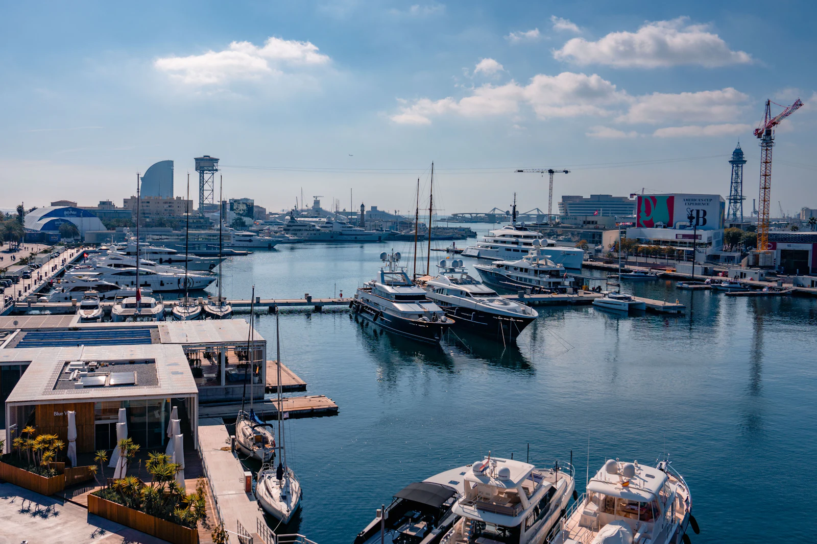 Superyachts and sailboats moored in Barcelona's Port Vell marina with cityscape and blue sky.