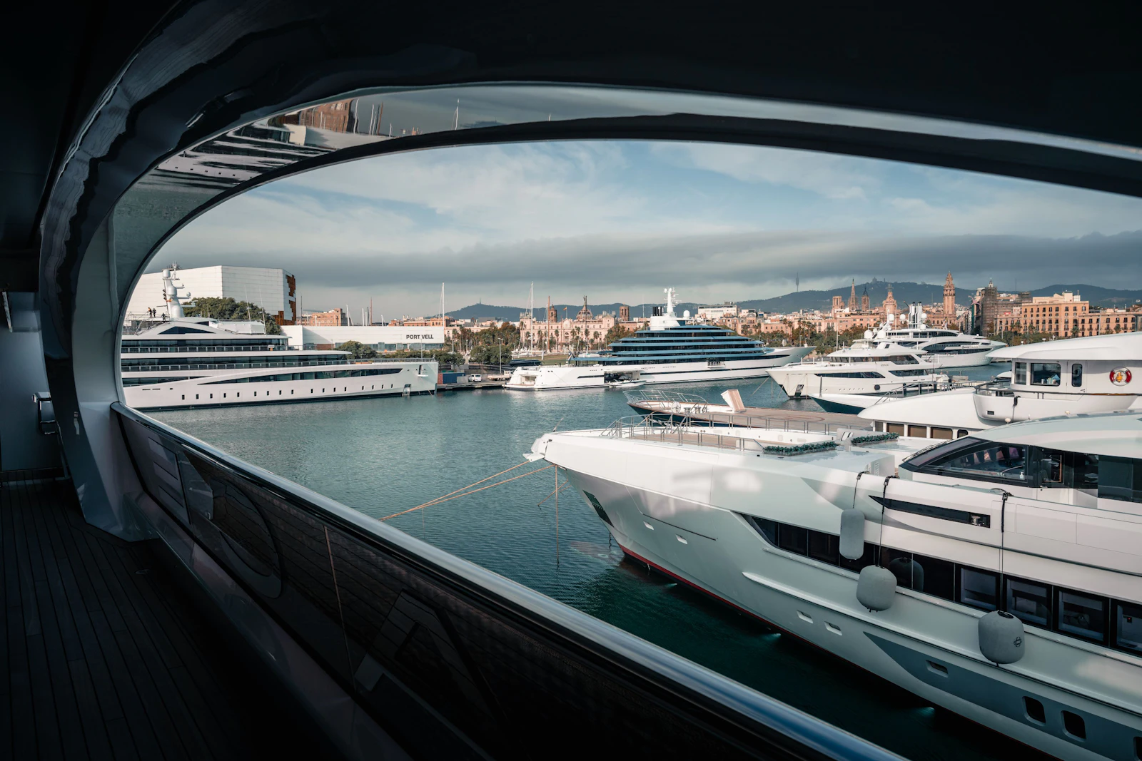 Vista a través de un ojo de buey de superyates amarrados en el puerto de Barcelona con el perfil de la ciudad y las montañas al fondo.