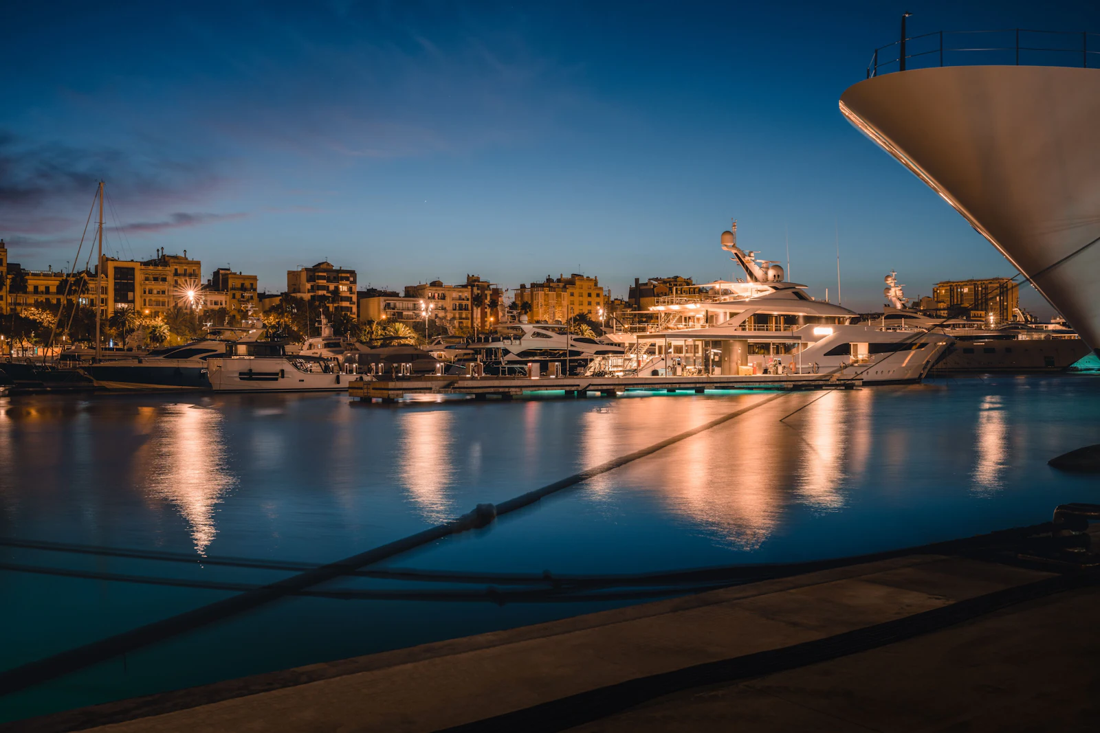 Superyachts moored at dusk in Barcelona's marina with illuminated city skyline reflected in calm water.