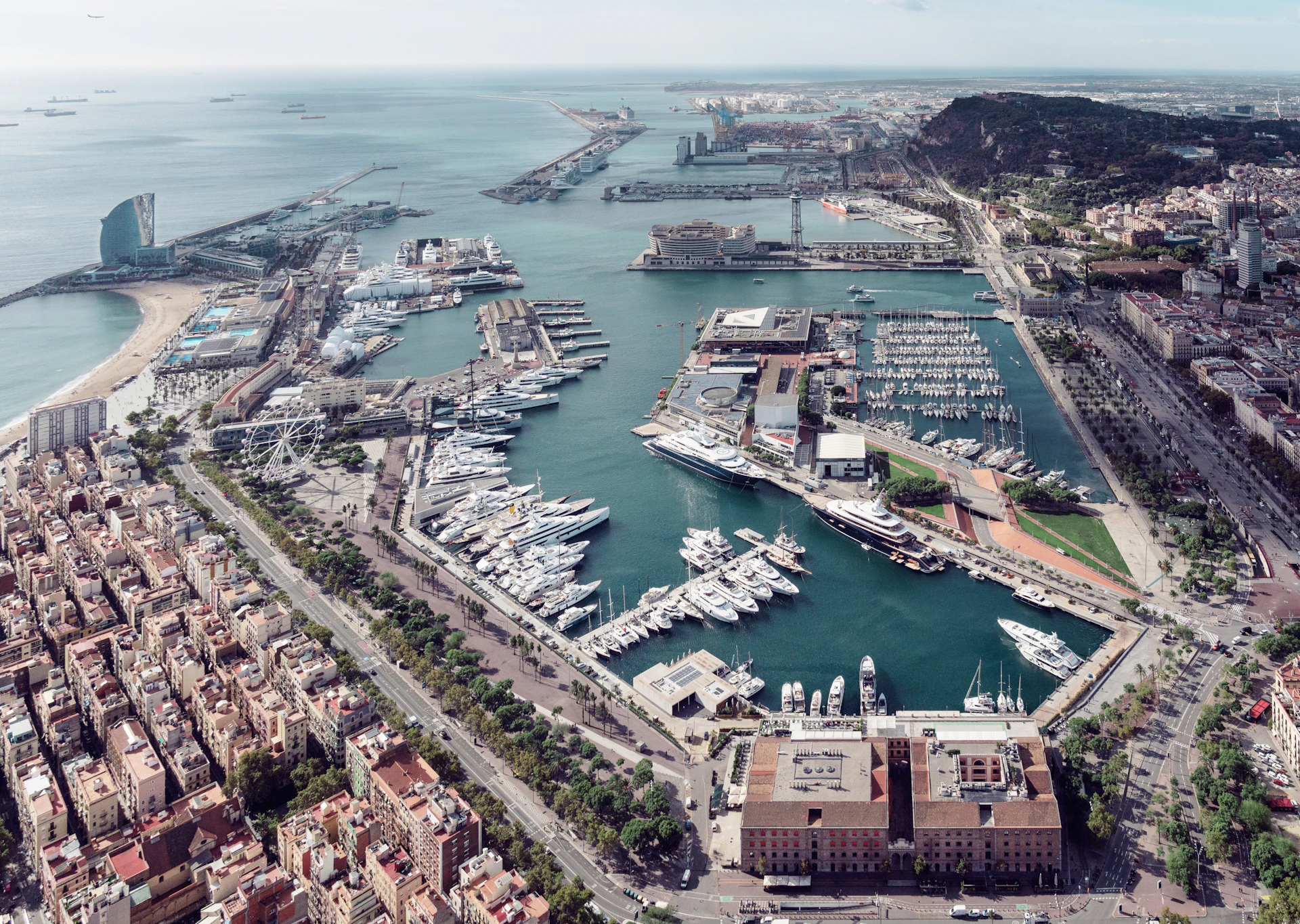 Aerial view of Barcelona's Port Vell marina with moored superyachts, surrounding waterfront buildings, and city extending to mountains in background.