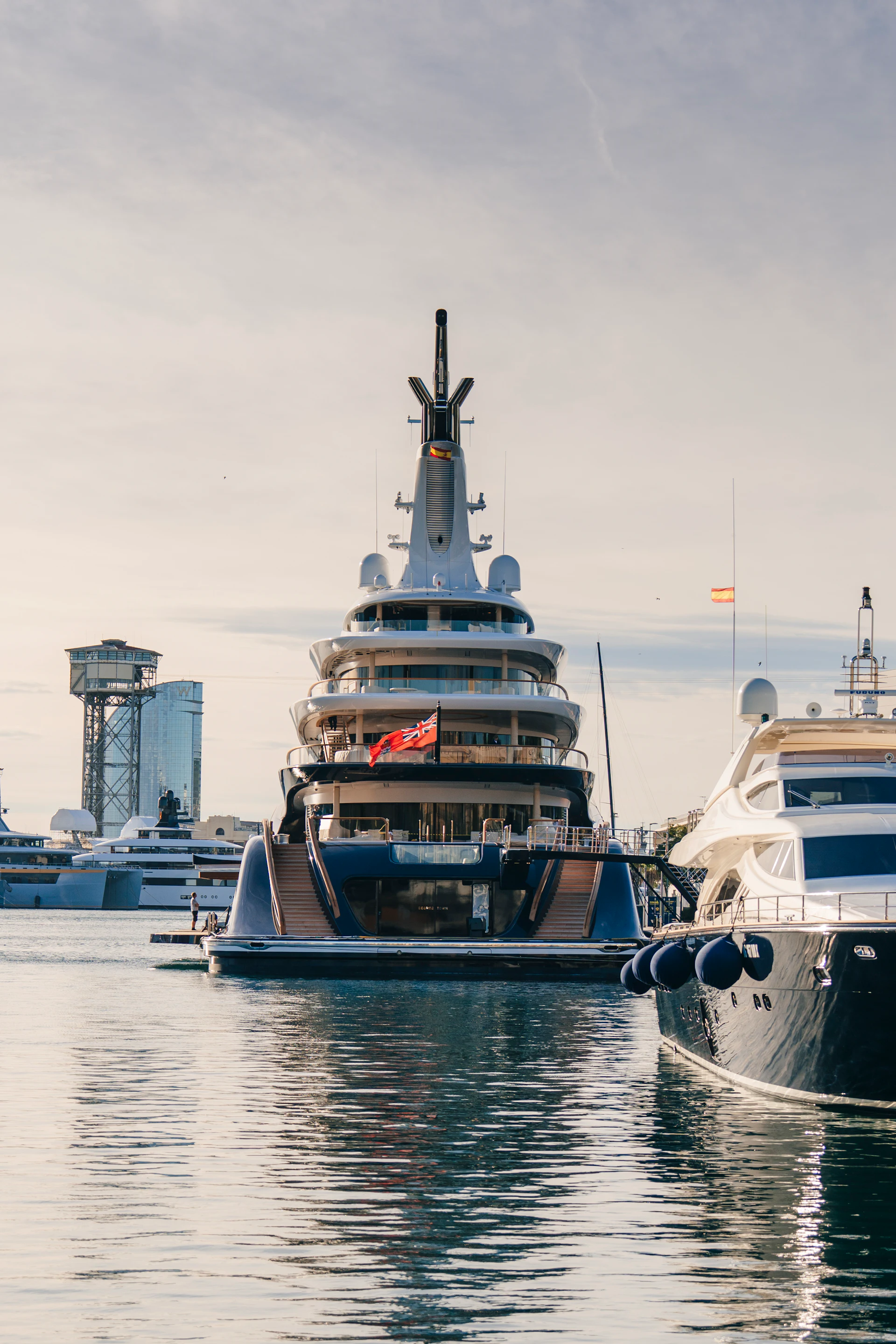 Large superyacht with multiple decks moored at marina alongside other vessels and waterfront structures.