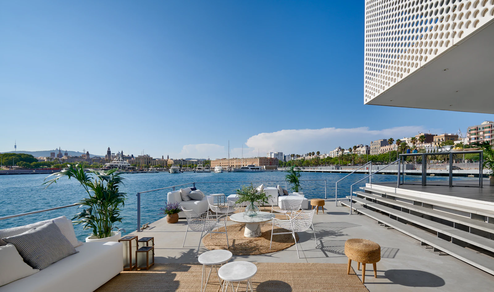 Terraza junto al mar con zonas de descanso con vistas al puerto y a la ciudad de Barcelona bajo un cielo azul despejado.