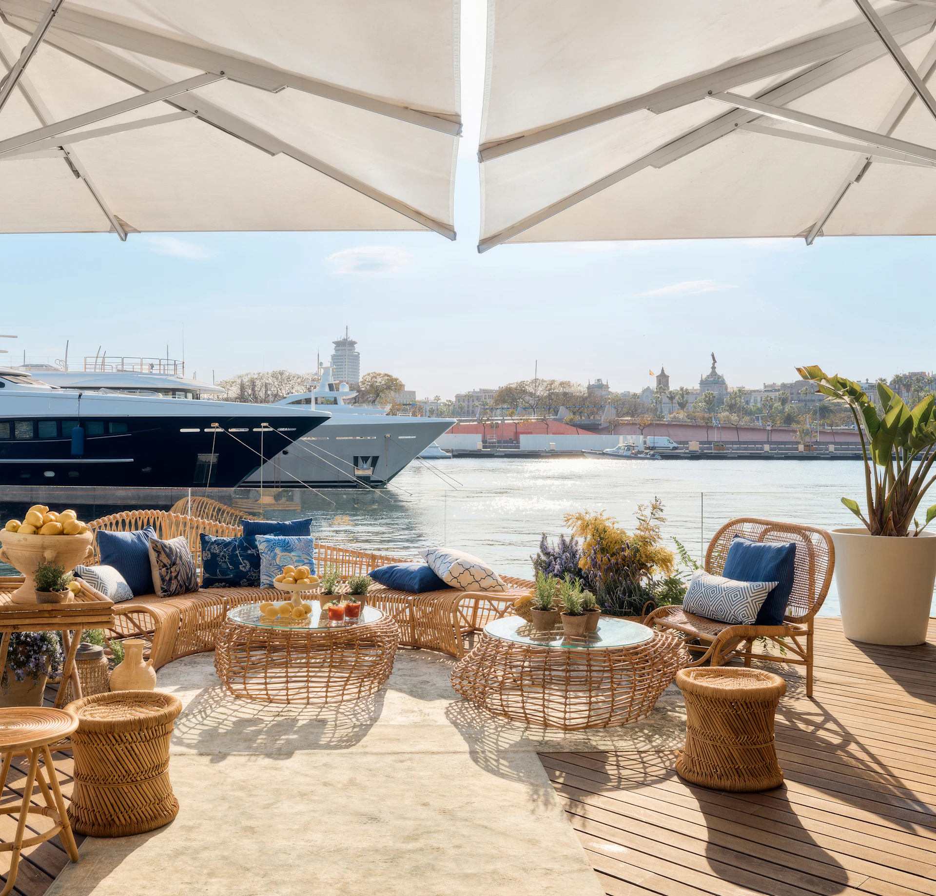 Waterfront terrace with wicker seating, white umbrellas, and Barcelona skyline across the marina.