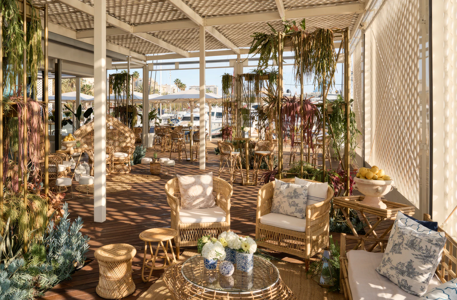 Sunlit covered terrace with wicker seating, hanging plants, and marina views in background.