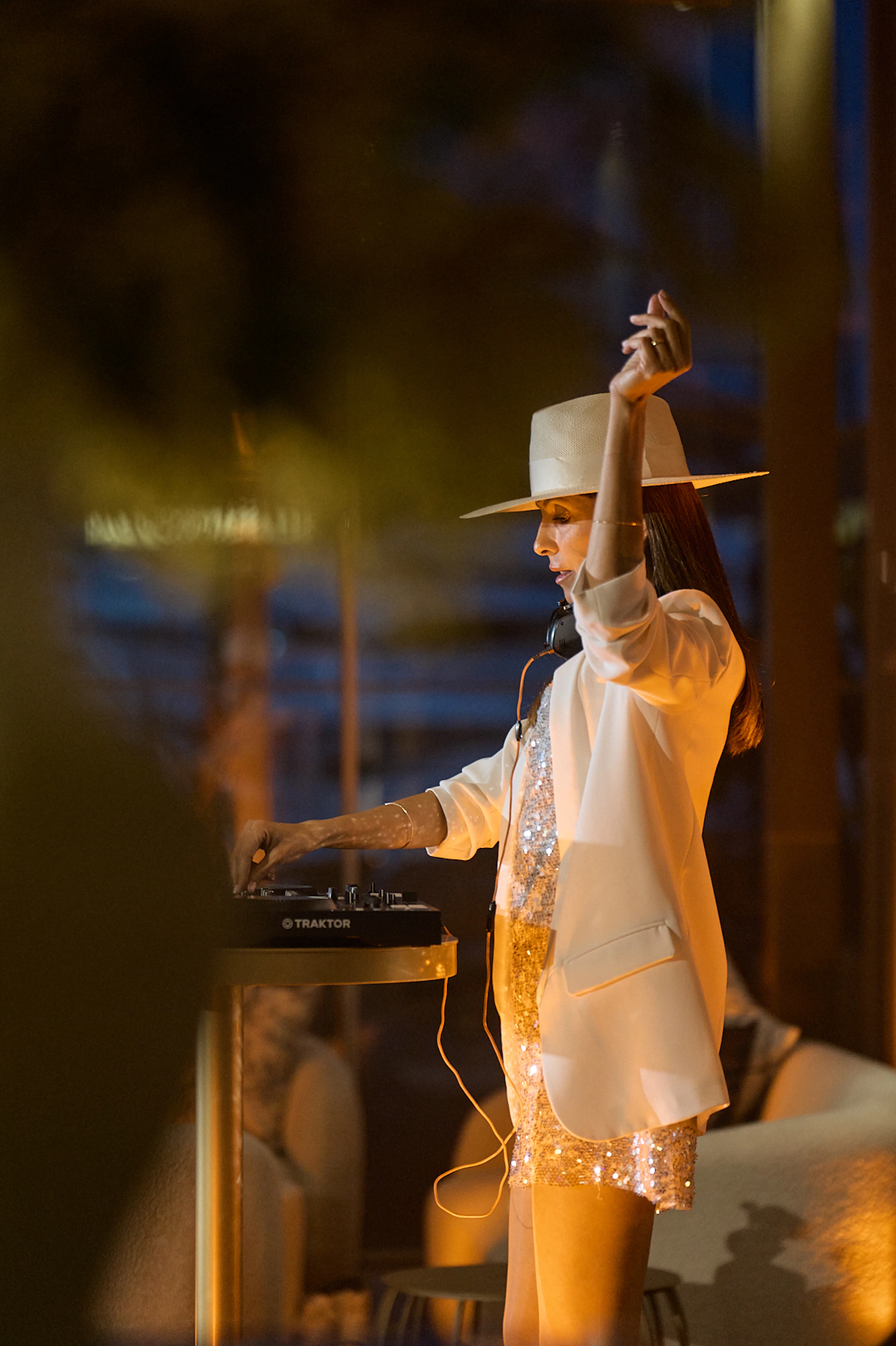 DJ in white hat and sequined outfit performing at a nighttime waterfront event.