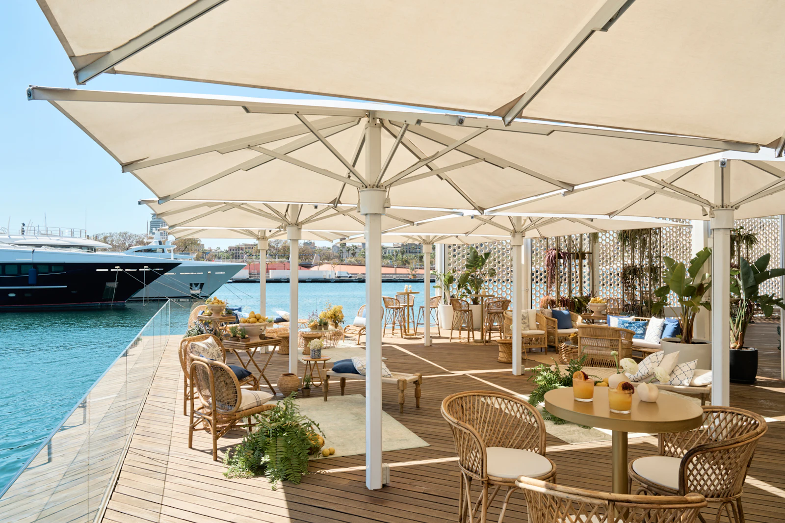 Terraza exterior junto al agua con grandes sombrillas blancas, mobiliario de ratán y superyates amarrados en el puerto deportivo.