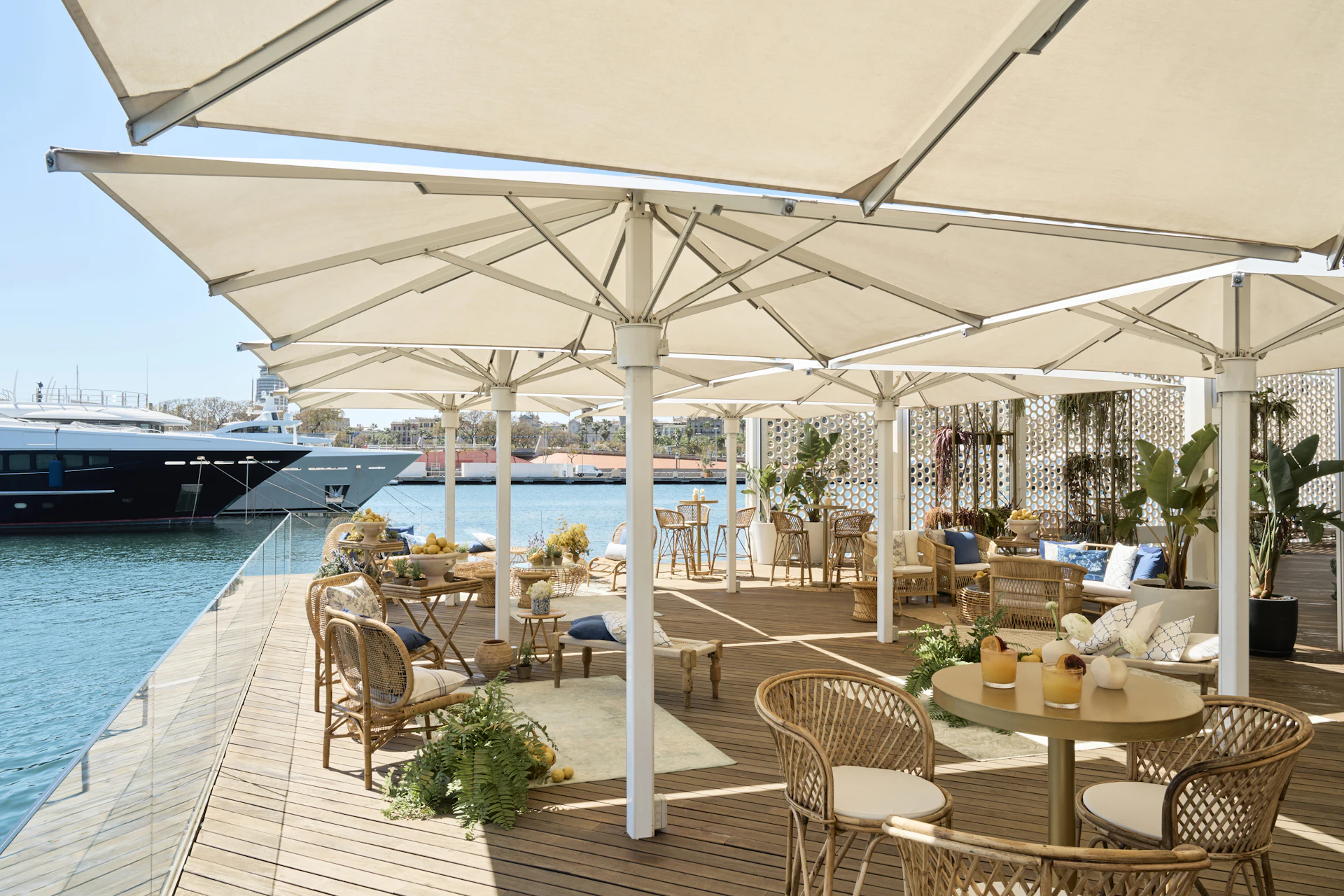 Waterfront terrace with rattan furniture and cream parasols overlooking moored superyachts.