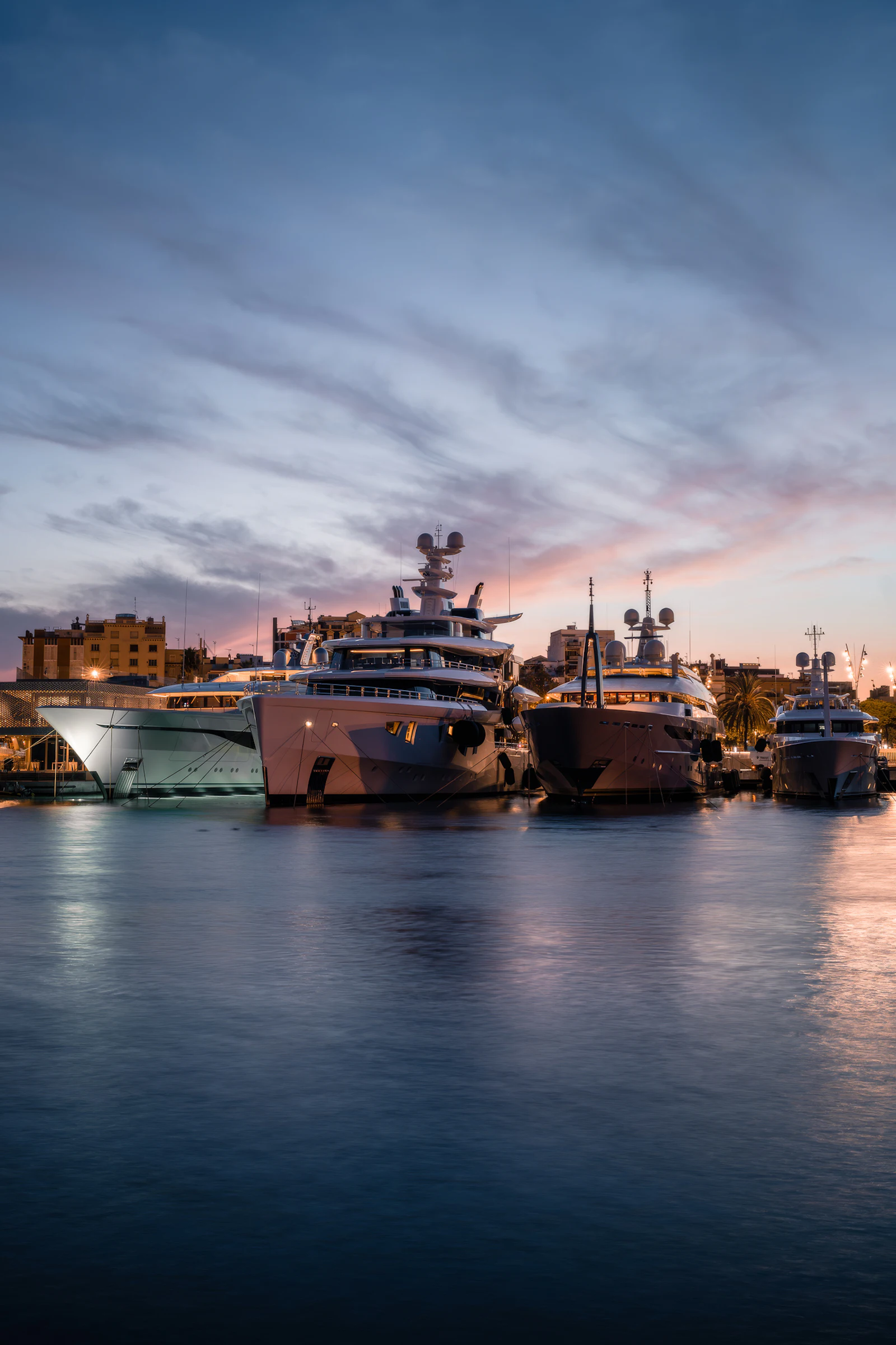 Multiple superyachts moored at sunset in a Mediterranean marina with Barcelona cityscape behind.