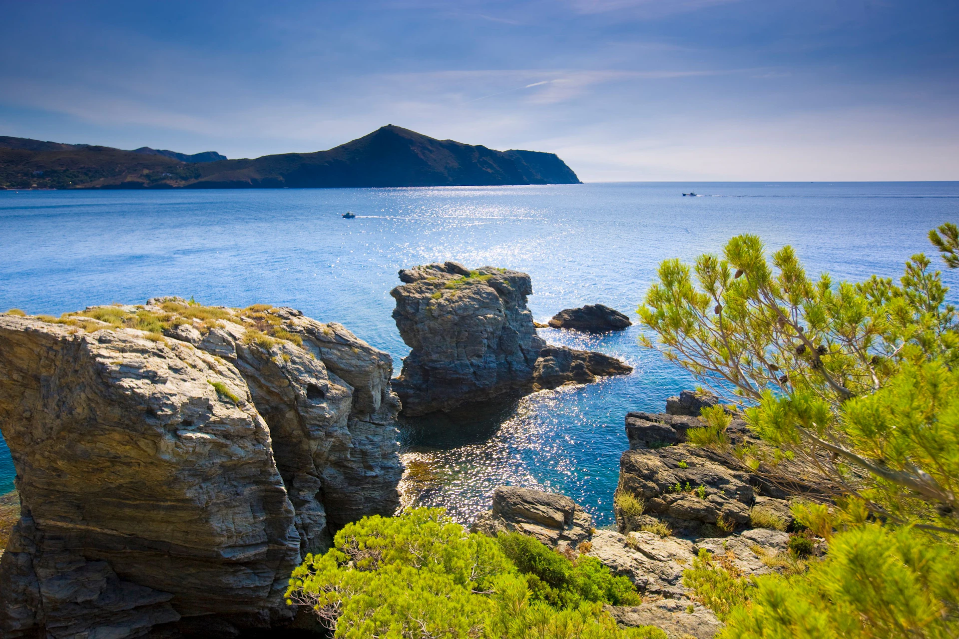 Rocky Mediterranean coastline with turquoise sea, distant island and moored boats.