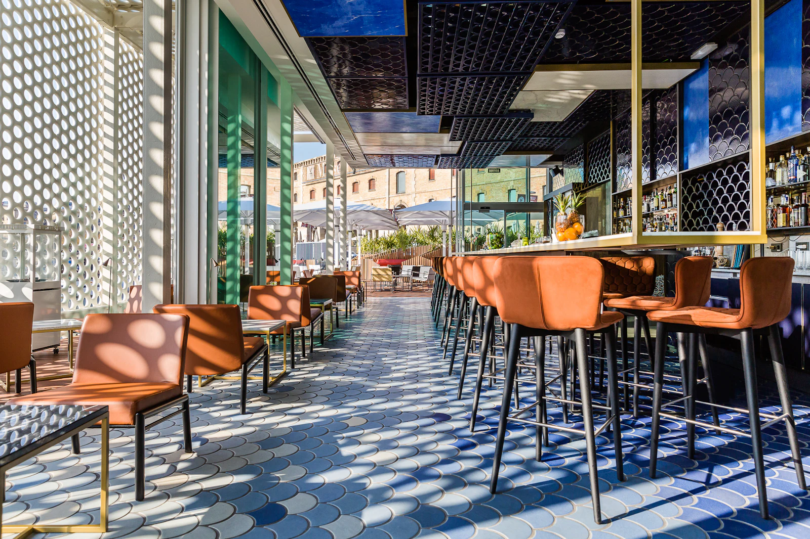 Interior de bar moderno con sillas naranjas, suelo estampado y vistas al paseo marítimo y los edificios de Barcelona.