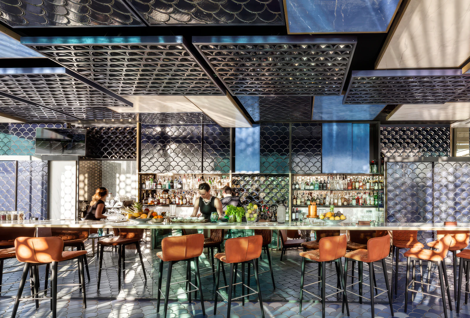Stylish bar with orange stools and blue fish-scale tiling, staff working behind the counter.