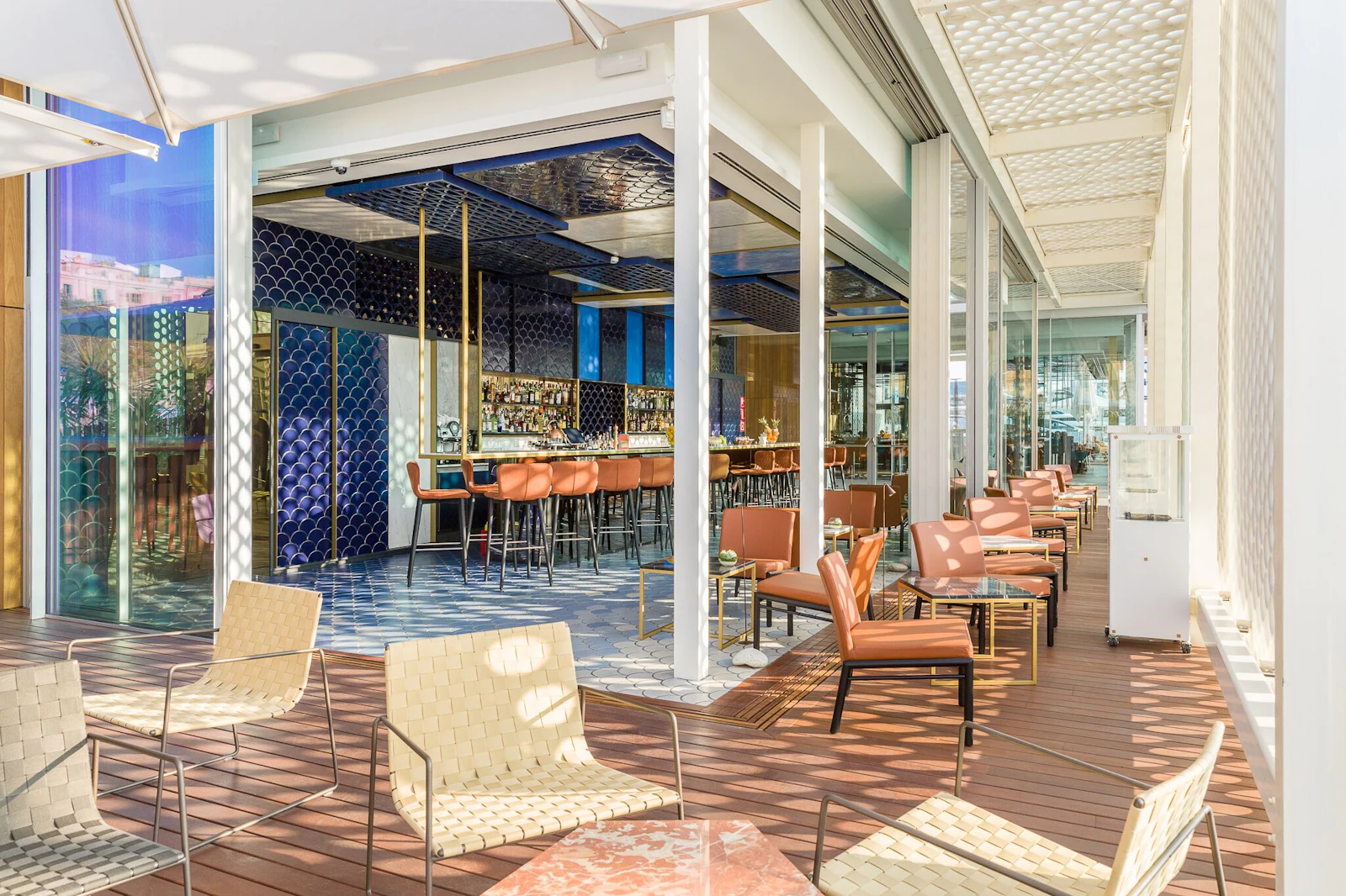 Modern waterfront lounge and bar with wooden deck seating, orange chairs, and blue interior design elements.