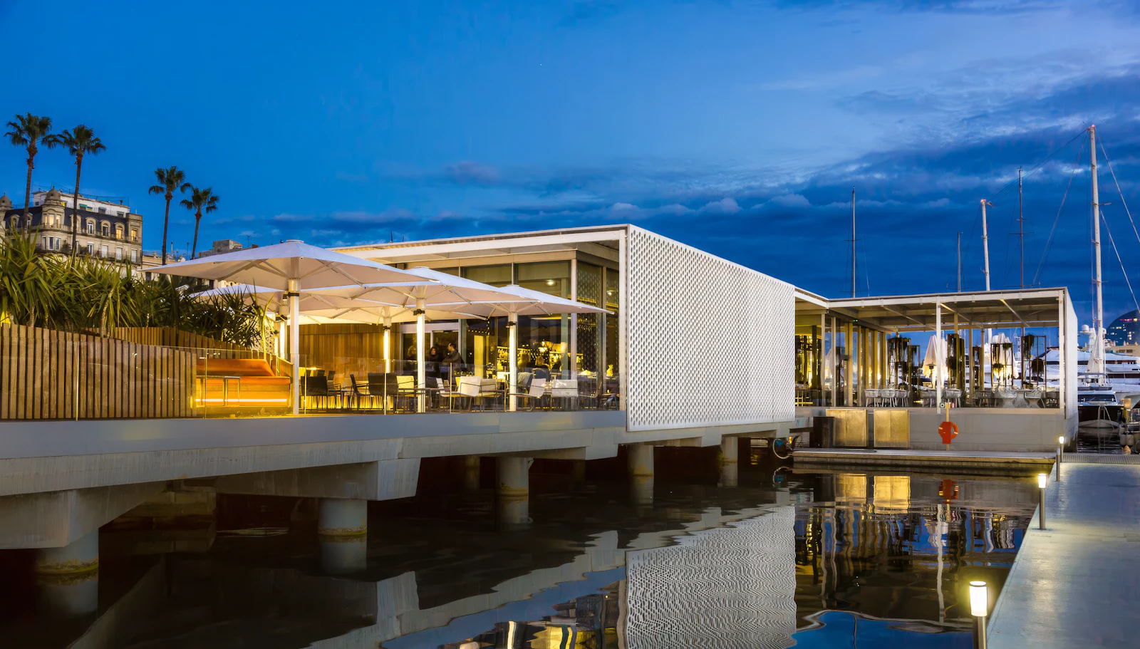 Restaurante moderno junto al agua con arquitectura blanca reflejada en el agua en calma al anochecer, con palmeras y yates amarrados visibles.