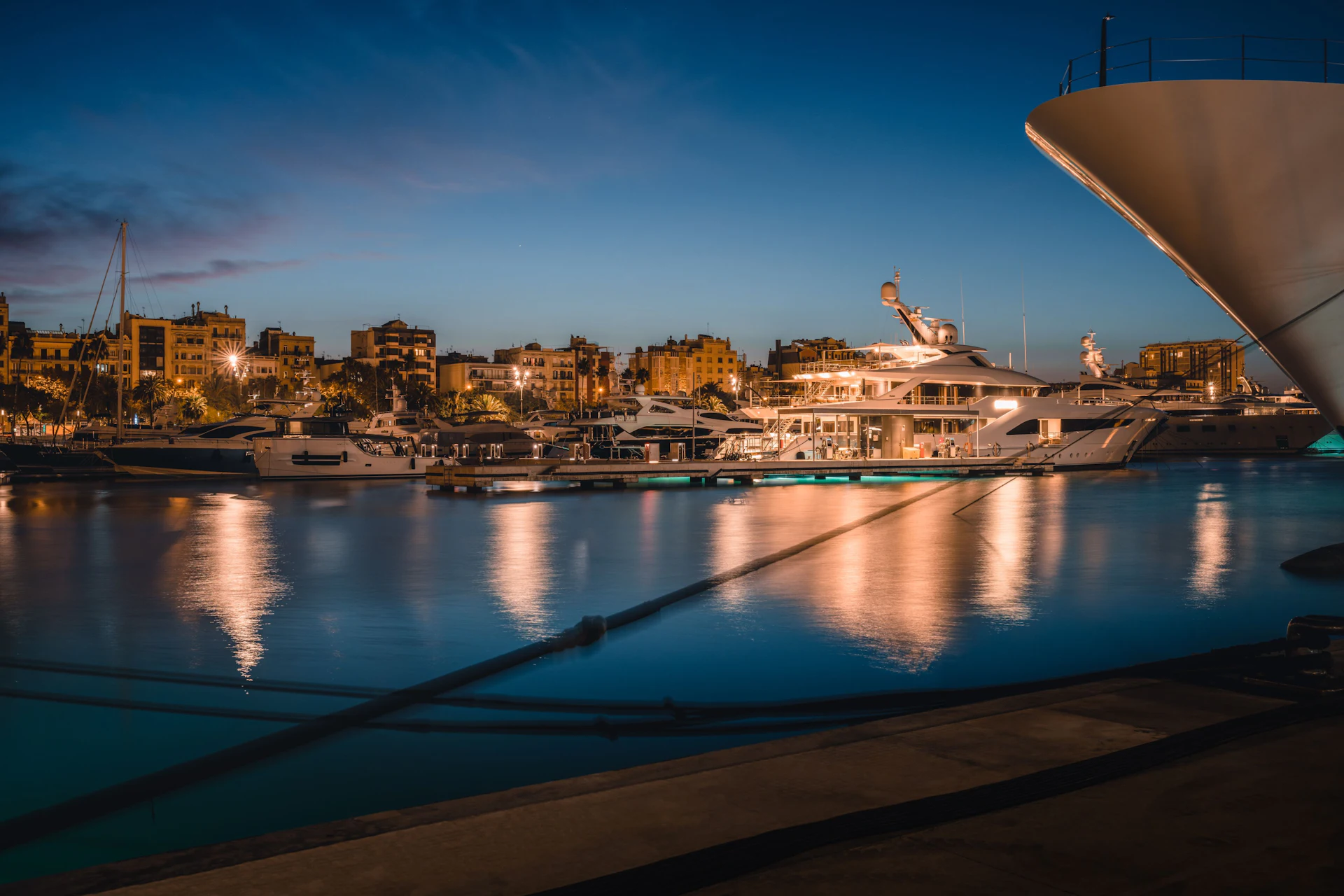 Superyates de lujo amarrados al atardecer en el puerto deportivo Port Vell de Barcelona con el perfil de la ciudad al fondo.