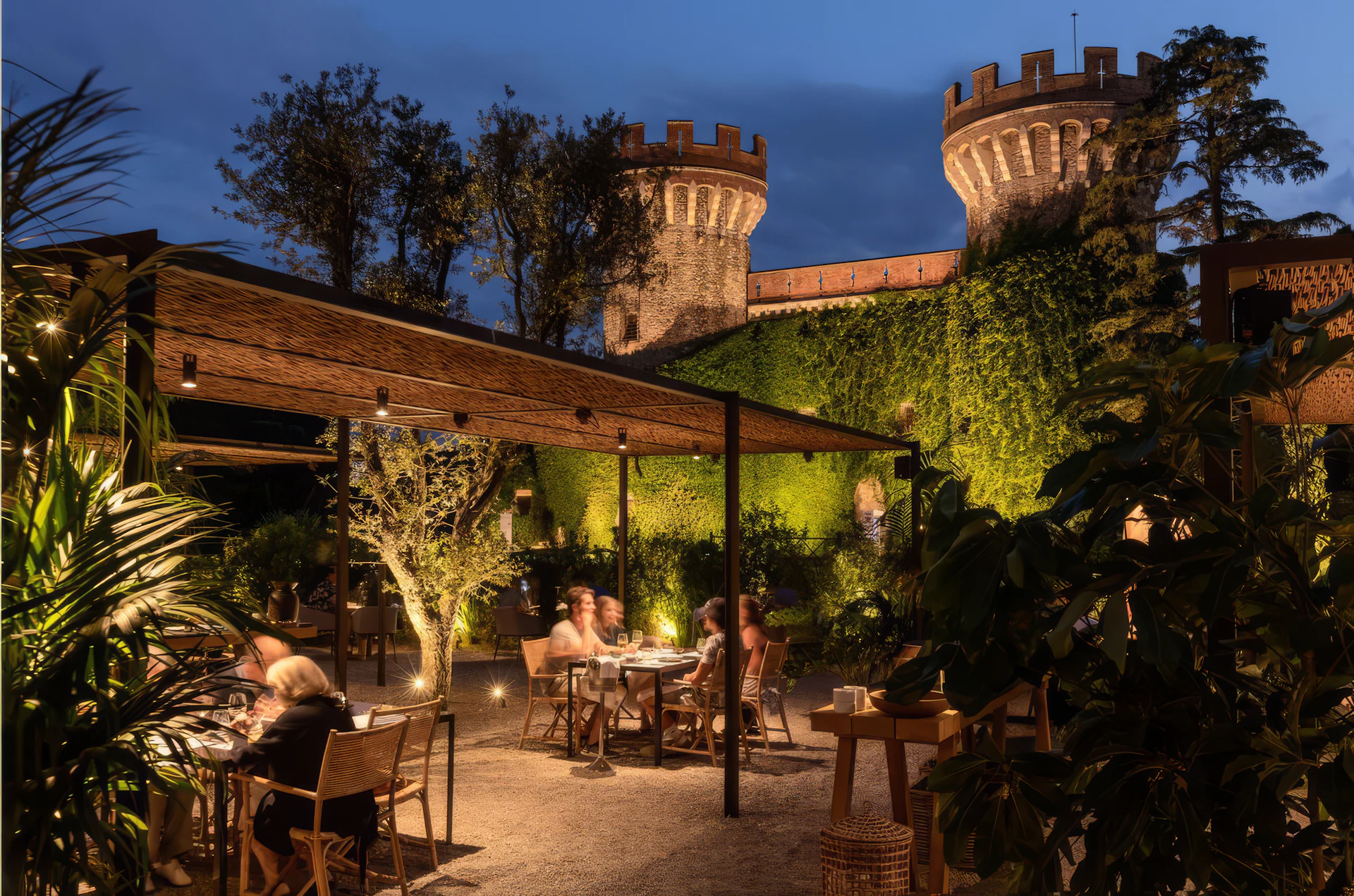 Patio iluminado con comensales bajo una pérgola de madera, rodeado de plantas tropicales y torres medievales de piedra al anochecer.