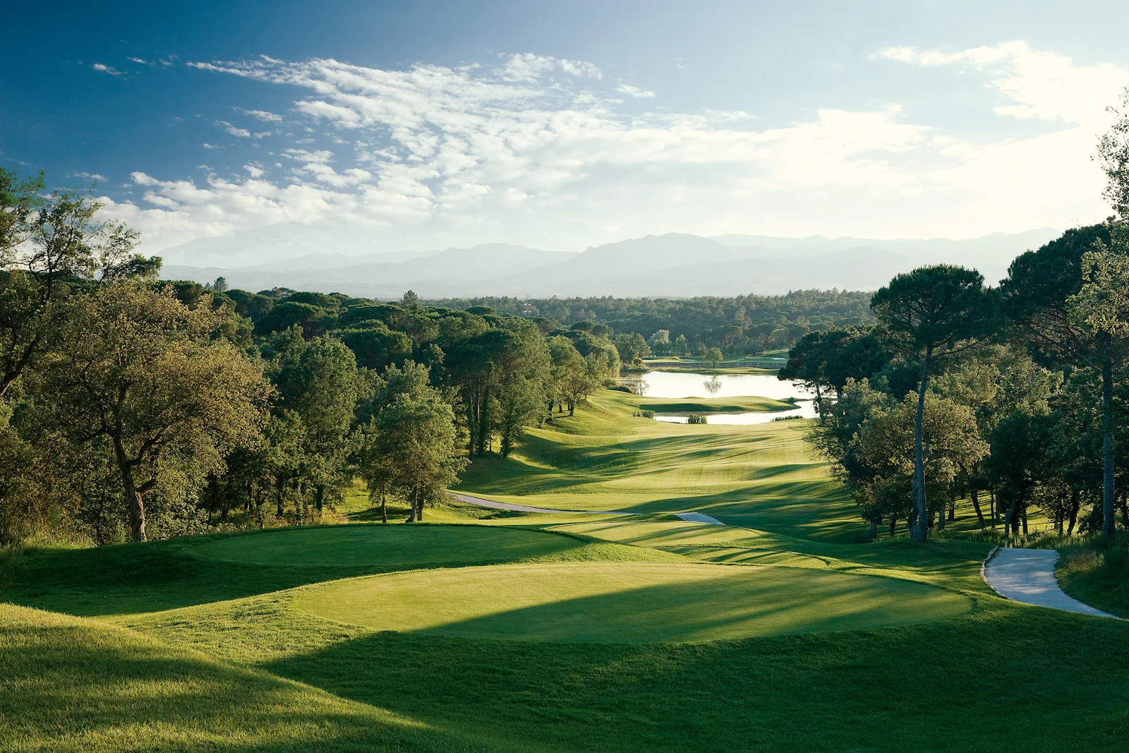 Golf course fairway with a lake, surrounded by trees and distant mountains.