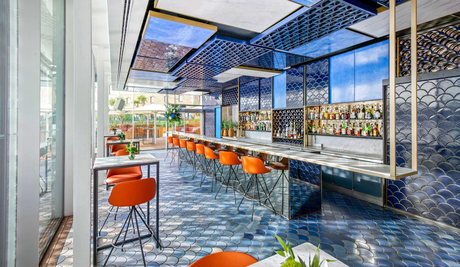 Zona de bar y lounge moderna con asientos naranjas, paneles de techo con estampado azul y vistas al puerto al fondo.