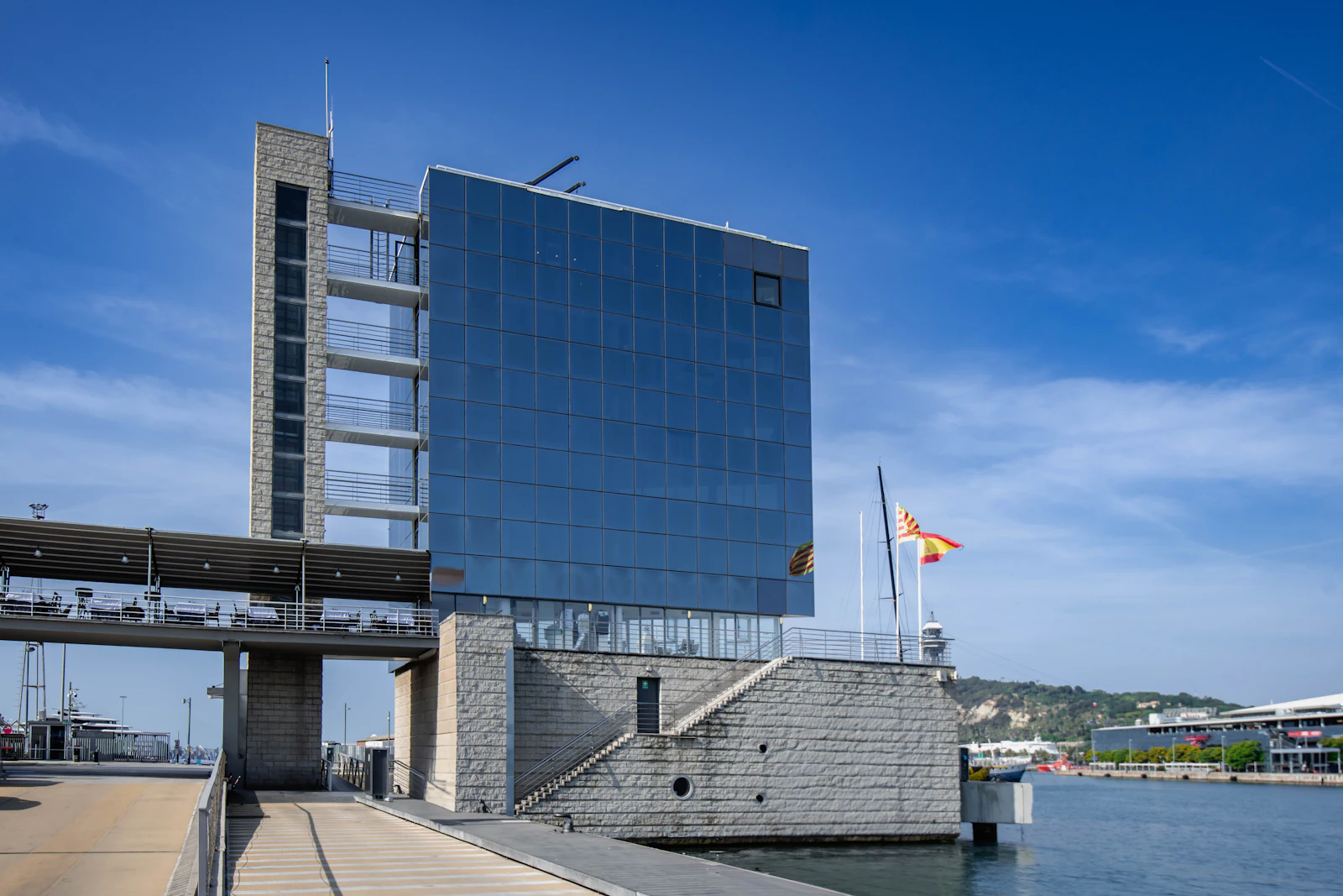 Modern waterfront building with blue-tinted glass facade beside Barcelona marina under clear sky.