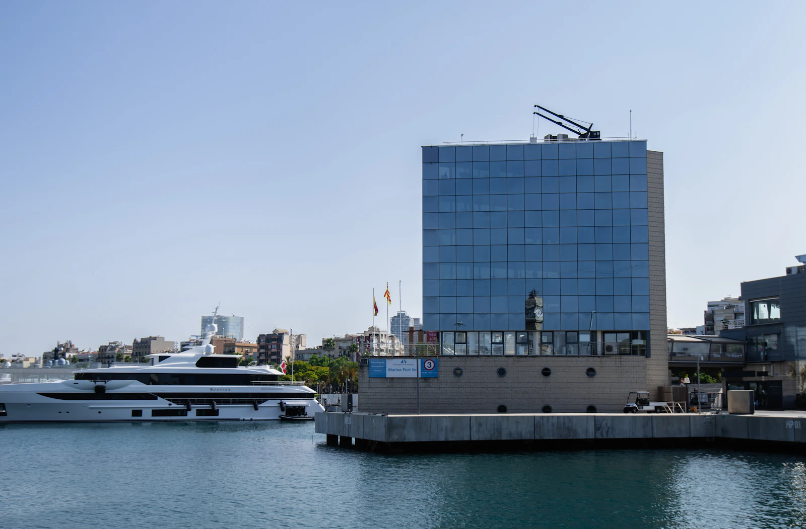 Superyate amarrado en Marina Port Vell con un edificio moderno frente al mar y el perfil urbano de Barcelona.
